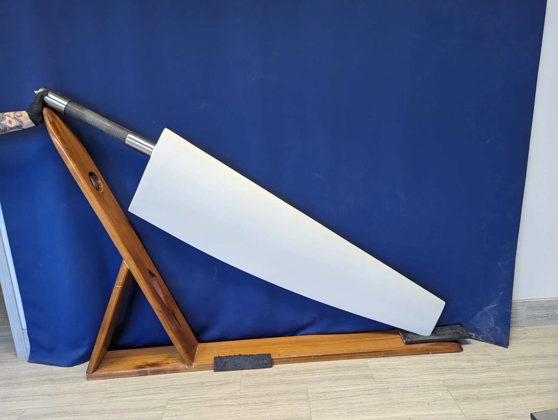 A person holding a large rudder with a wooden handle and a wide blade. The background is a blue curtain, and part of a wooden stand or support is visible.