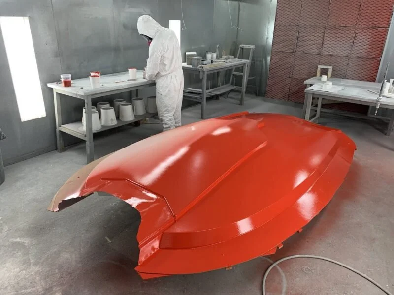 A person in protective clothing working on a glossy red boat shell inside a workshop.