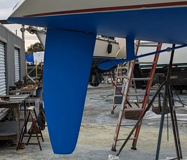 Close-up of a boat's blue keel, with workbenches, ladders, and boatyard equipment in the background, indicating maintenance or construction work.