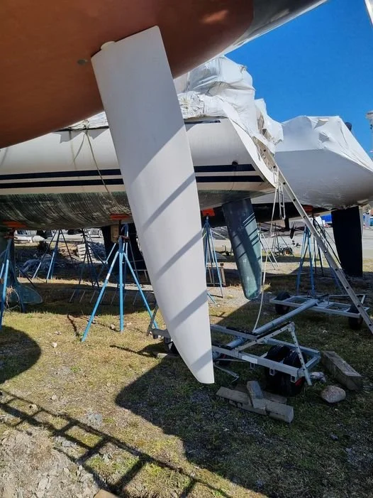 A sailboat on land supported by multiple blue stands, with a white keel and hull, and a partially covered deck, in a boatyard under a clear blue sky.