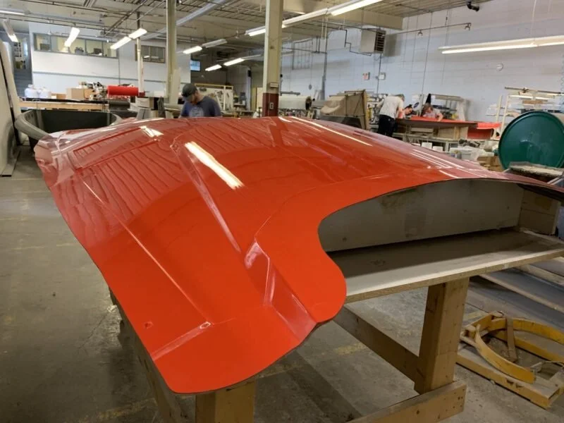 A red boat hull is being assembled on a workbench in a factory. Workers and tools are visible in the background.