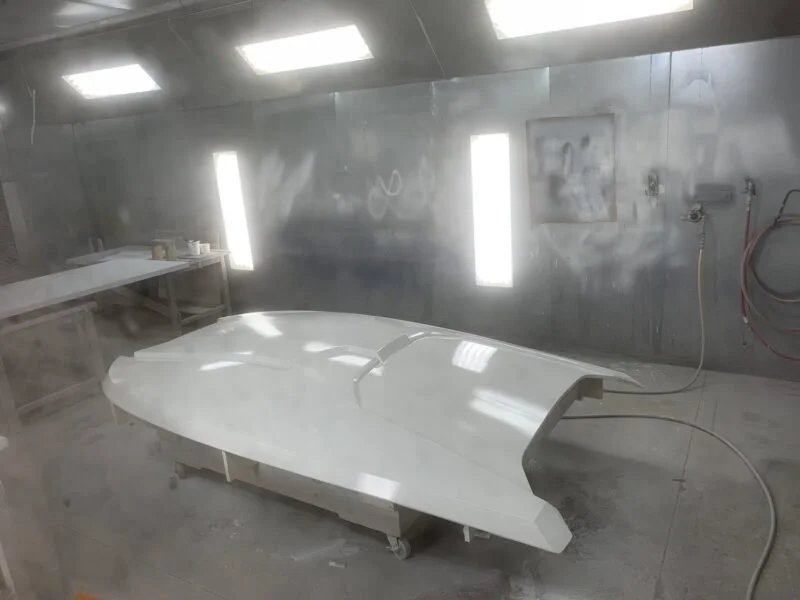 A sleek, boat hull on a work cart inside a paint booth, with bright overhead lights and a stainless steel wall in the background.
