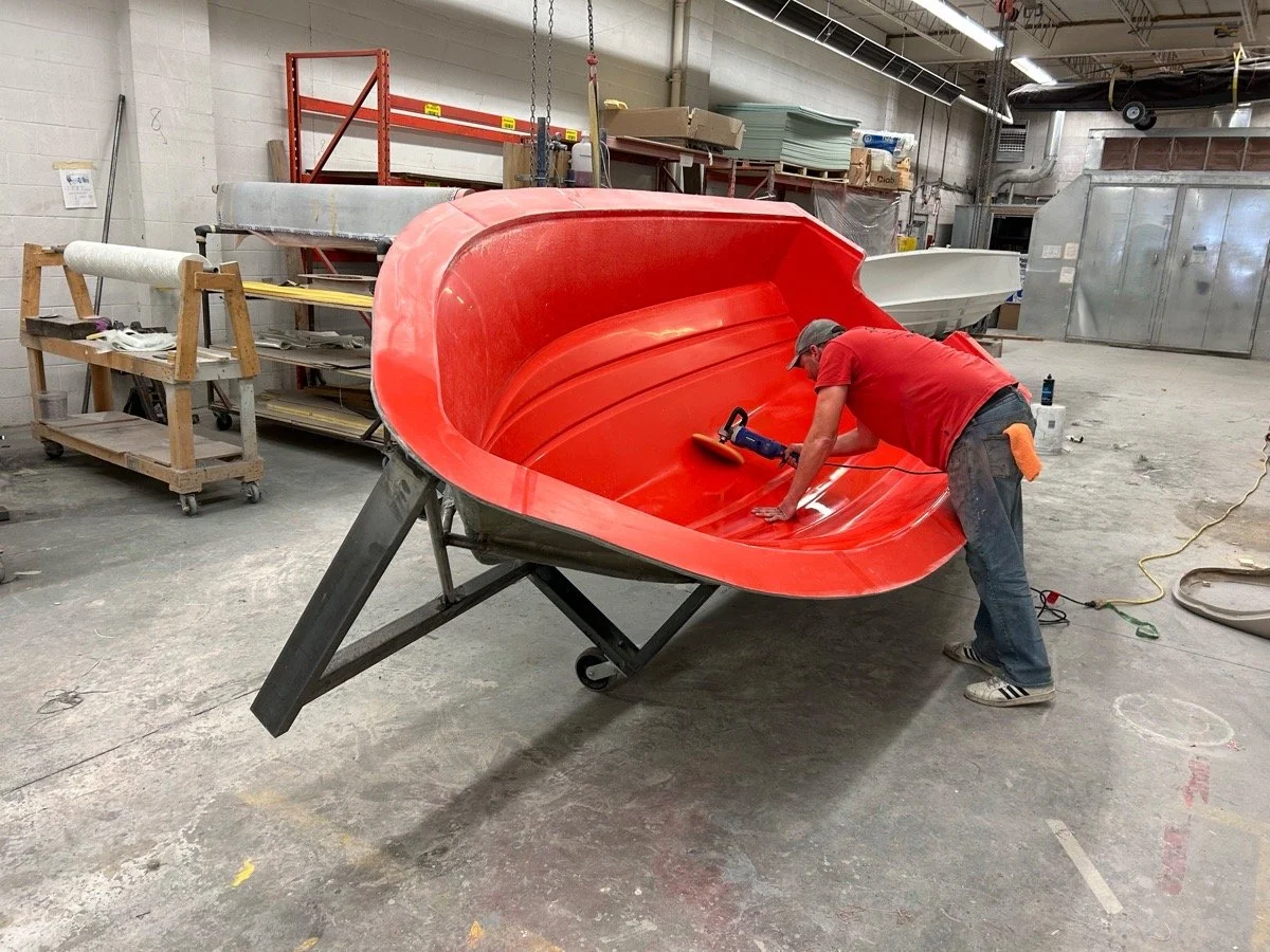A worker in a workshop is painting a red boat hull with a spray gun. The boat is positioned on a support stand, and the workspace contains tools, shelves, and equipment.