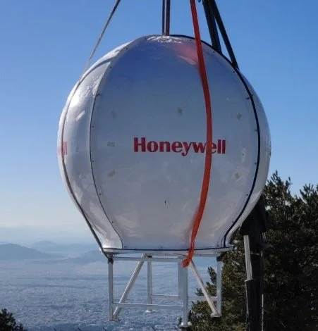 Large white spherical weather radar antenna with the Honeywell logo, mounted on a metal frame outdoors with a view of a body of water and distant mountains.