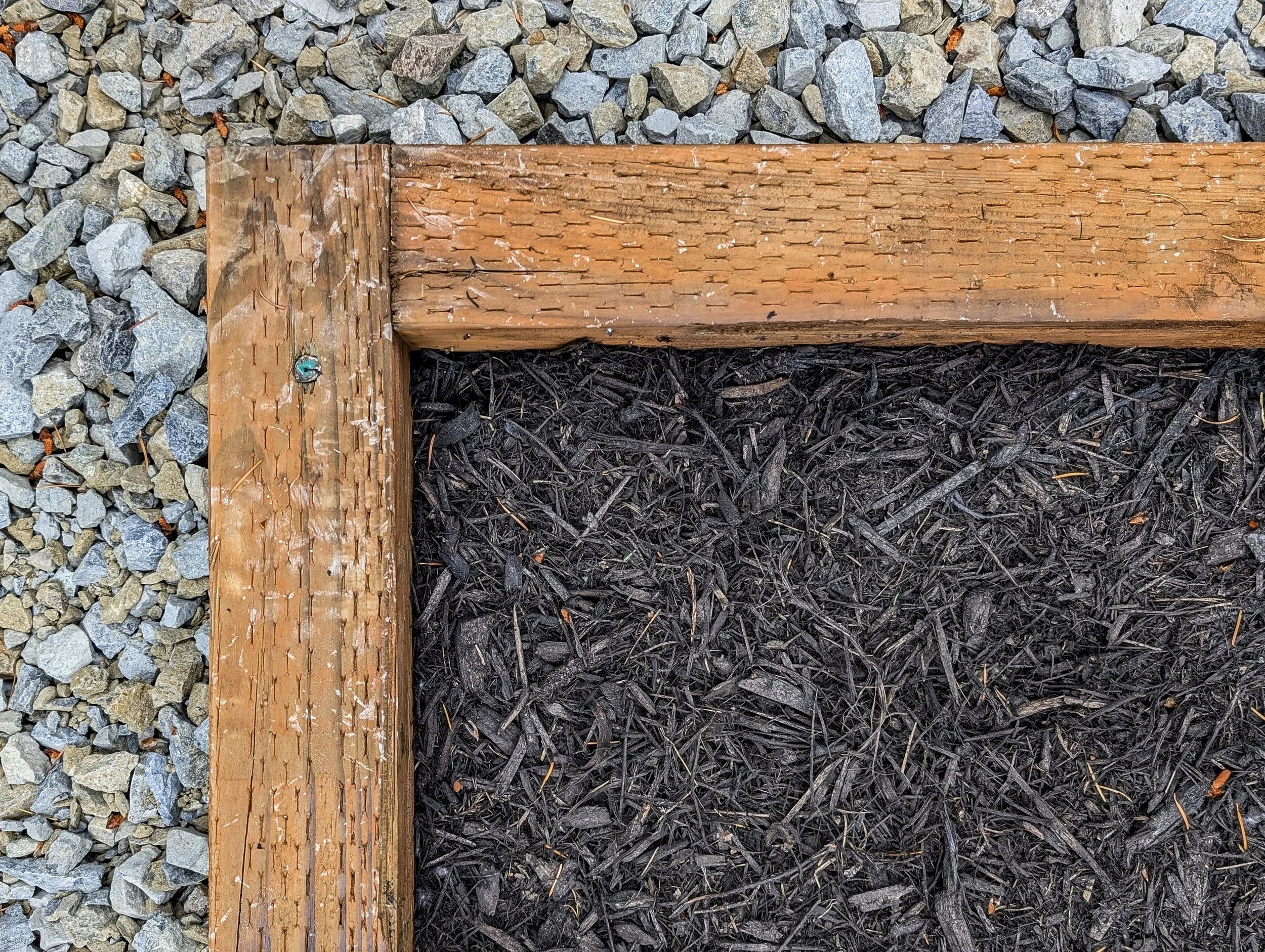 Close-up of a wooden tent pad corner with gravel on one side and dark soil with mulch inside.