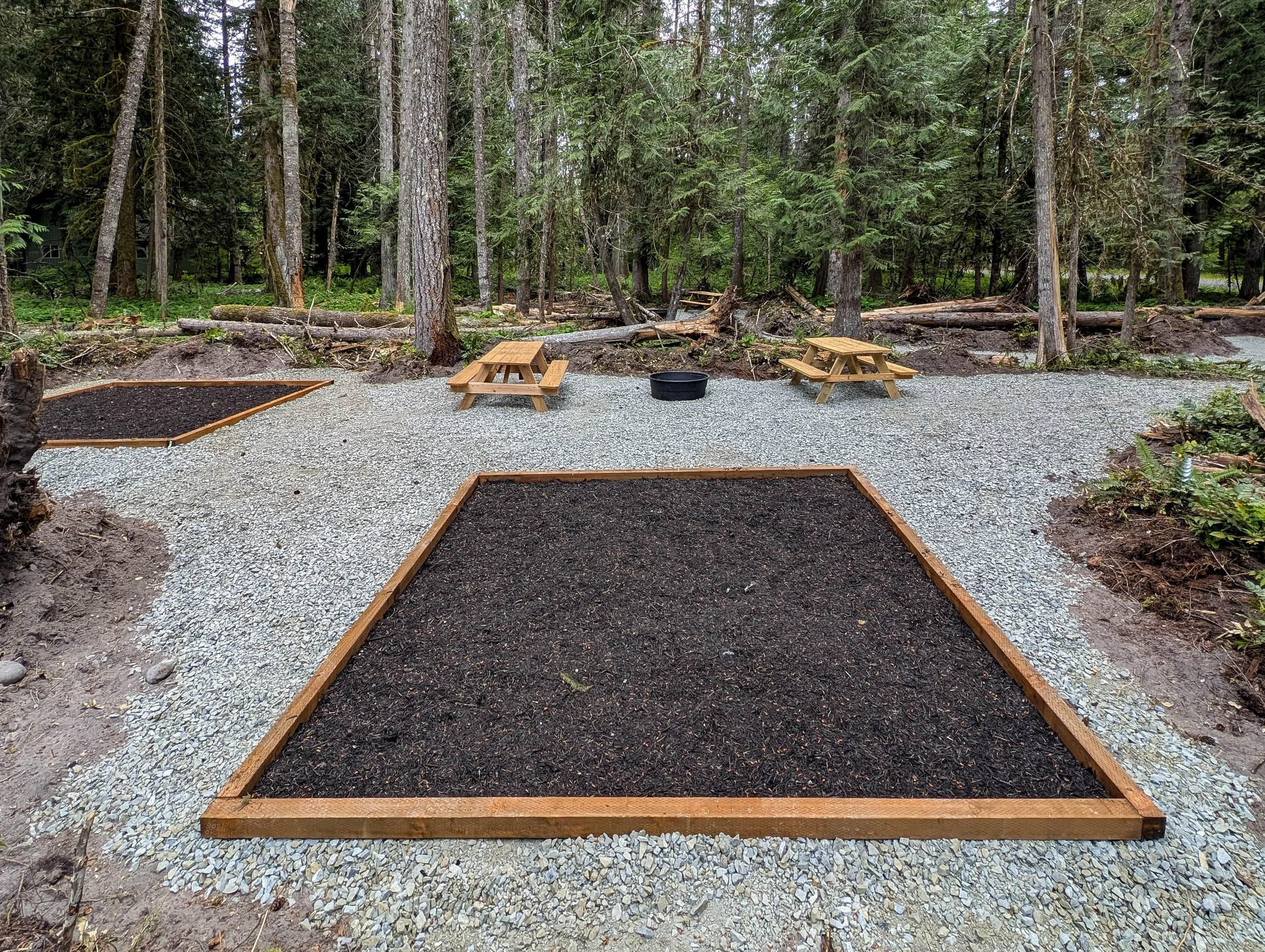 Two tent pads on a Mulch bed surrounded by gravel with two small wooden picnic tables and a black fire pit in a forested area.