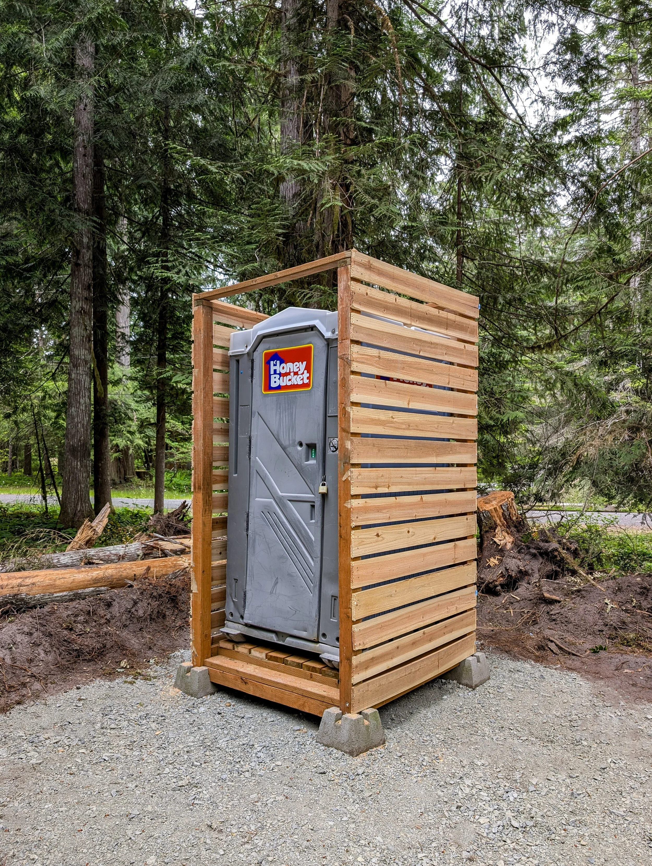 Portable toilet enclosed in a wooden frame with slatted walls in a forest setting.