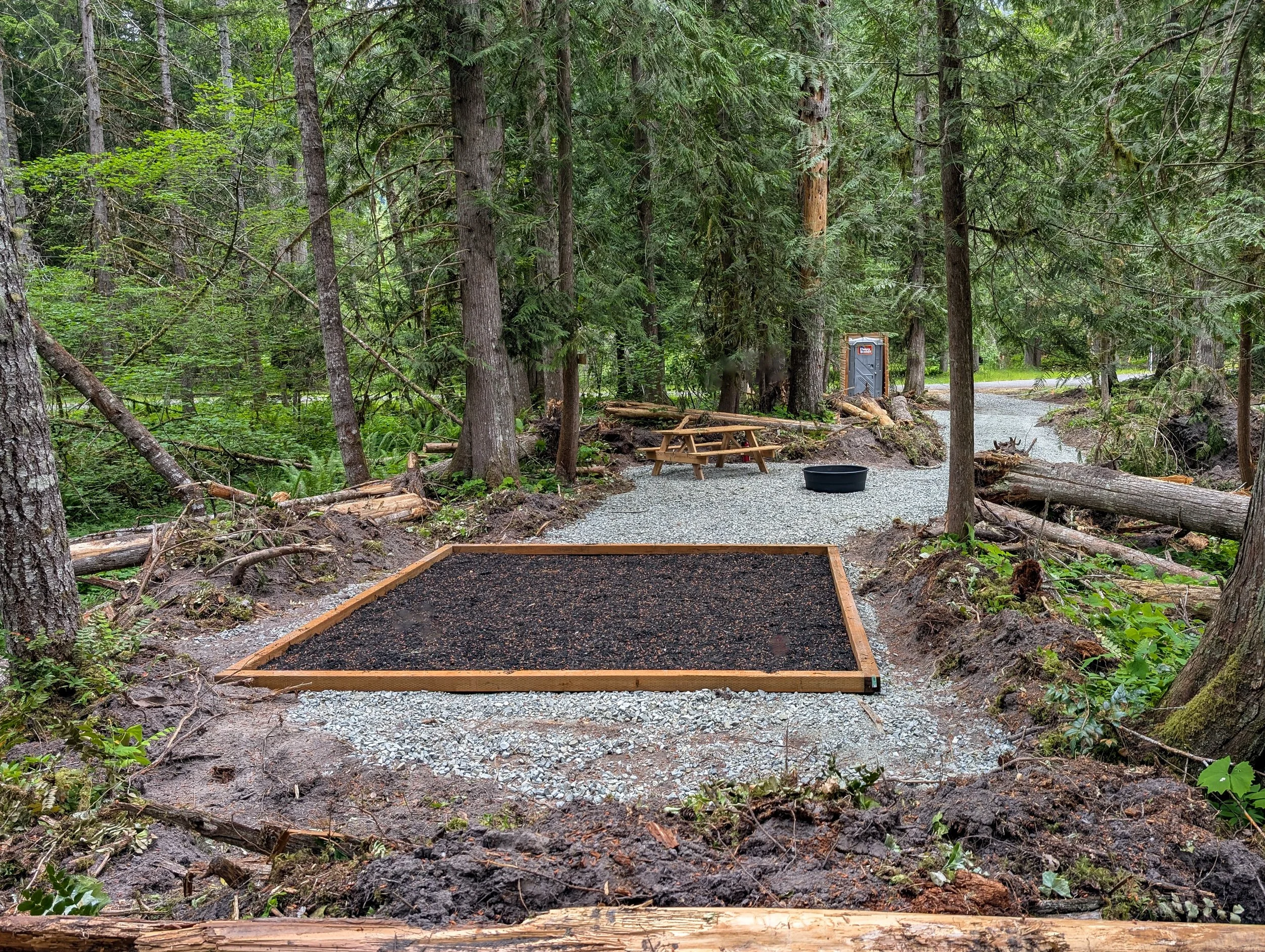 A forest scene with a gravel pathway, a small wooden picnic table, a portable toilet, and a patch of black mulch bordered by a wooden frame for a tent pad, surrounded by tall trees and fallen logs.