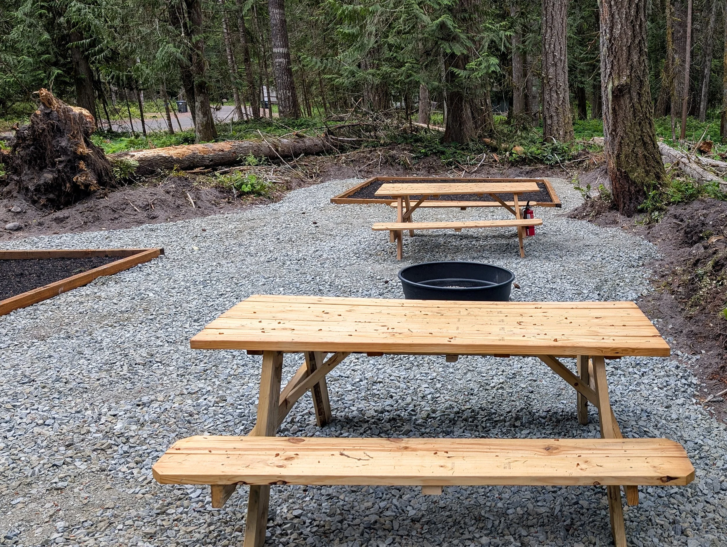 Open outdoor campsite with two wooden picnic tables, a black fire pit, and a gravel surface surrounded by trees.
