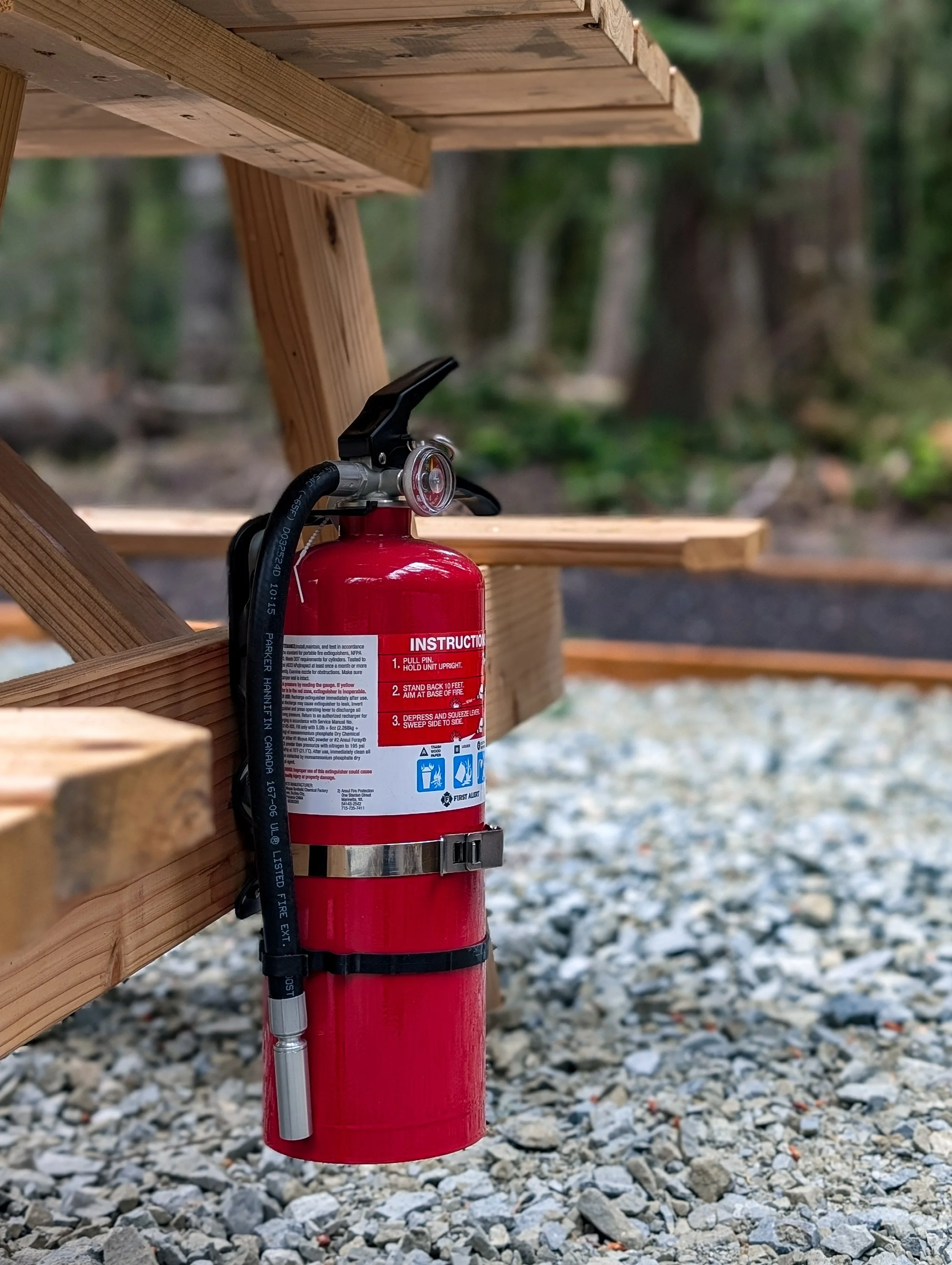 A red fire extinguisher attached to a wooden picnic table outdoors with trees and gravel in the background.