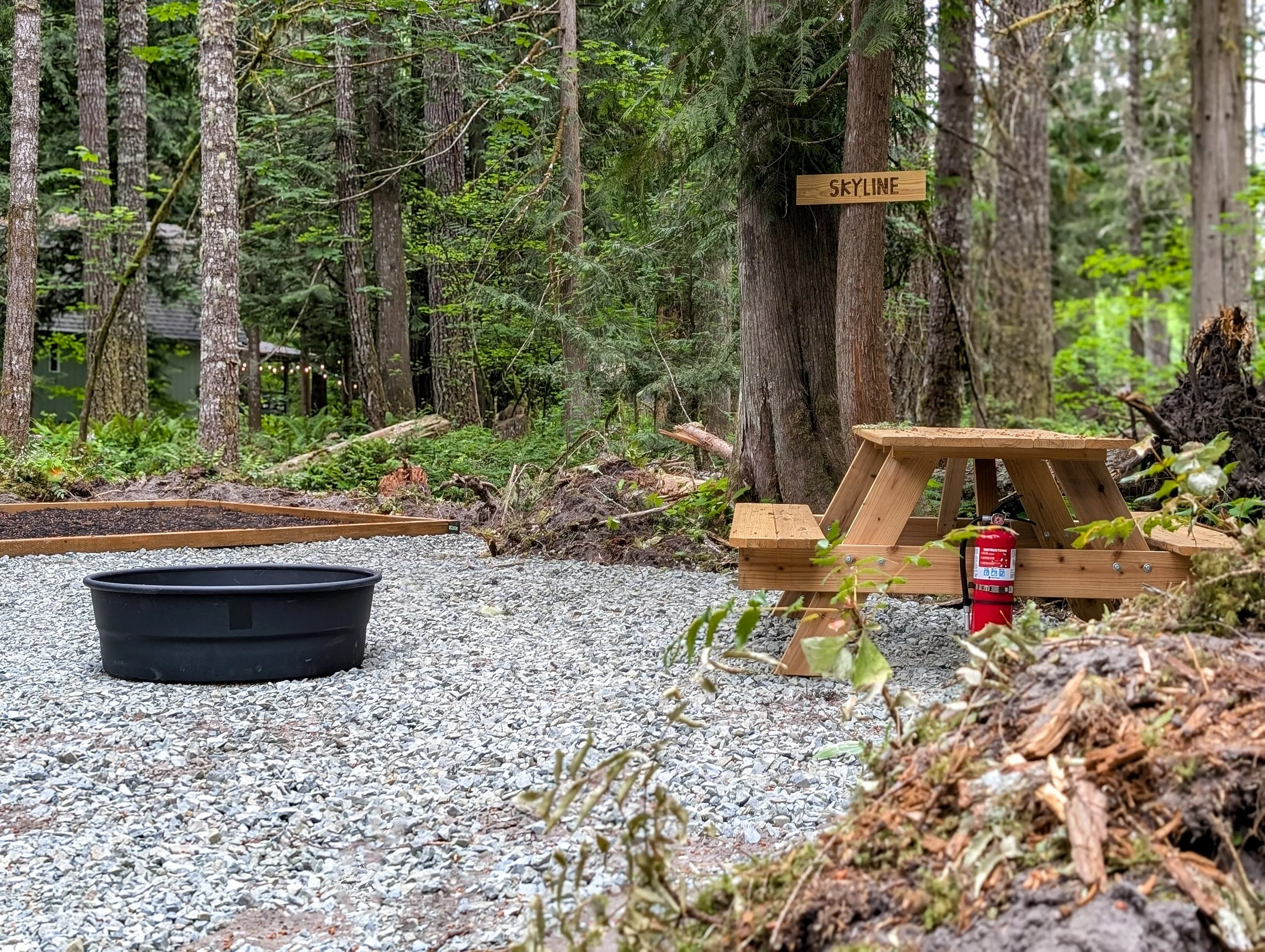 A forest scene with a small black fire pit, a wooden picnic table, and a fire extinguisher. There is a sign on a tree that reads 'Skyline.' The area is covered with gravel and surrounded by tall trees and green foliage.