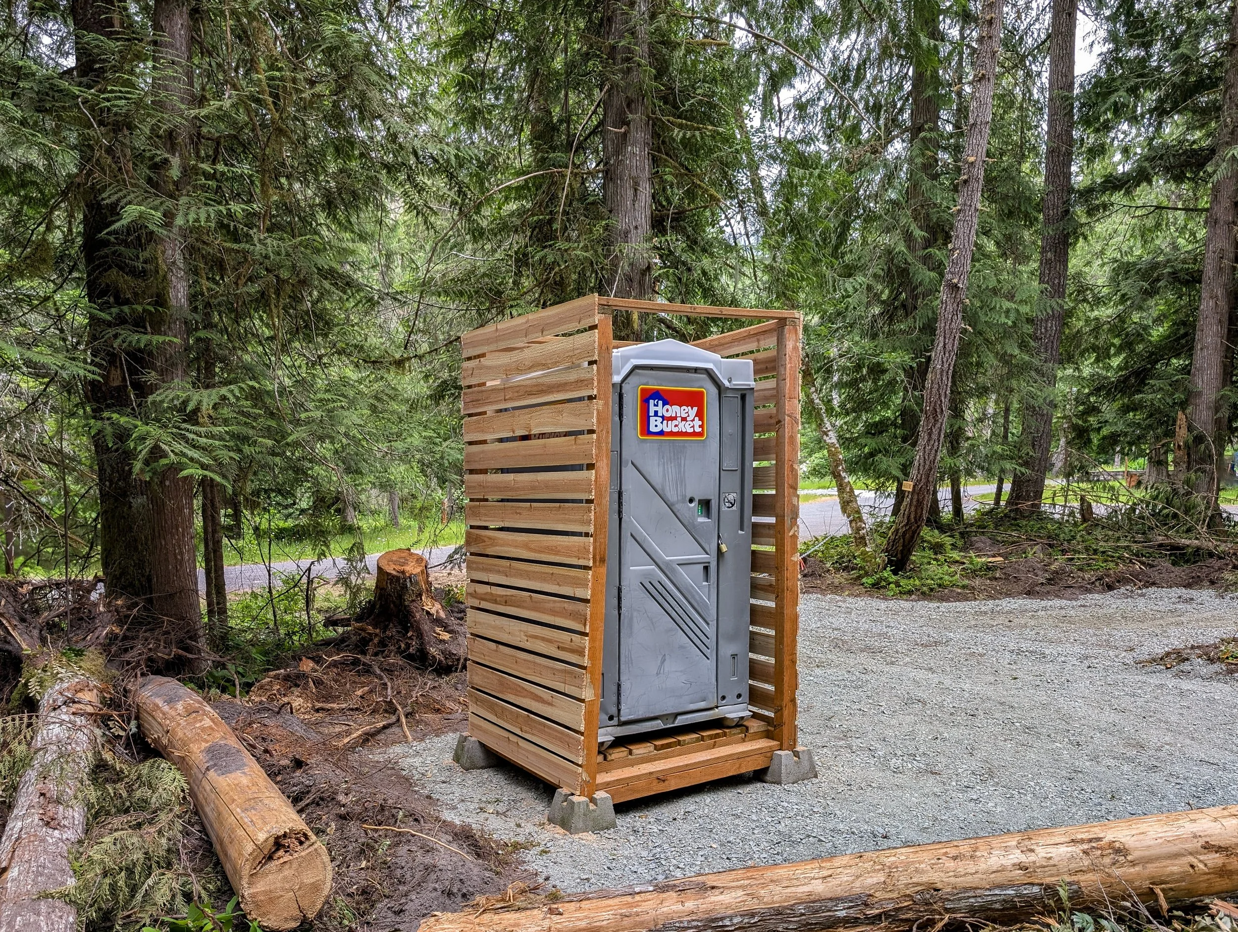 Portable toilet labeled 'Honey Bucket' inside a wooden enclosure in a forested area with trees and logs on the ground.