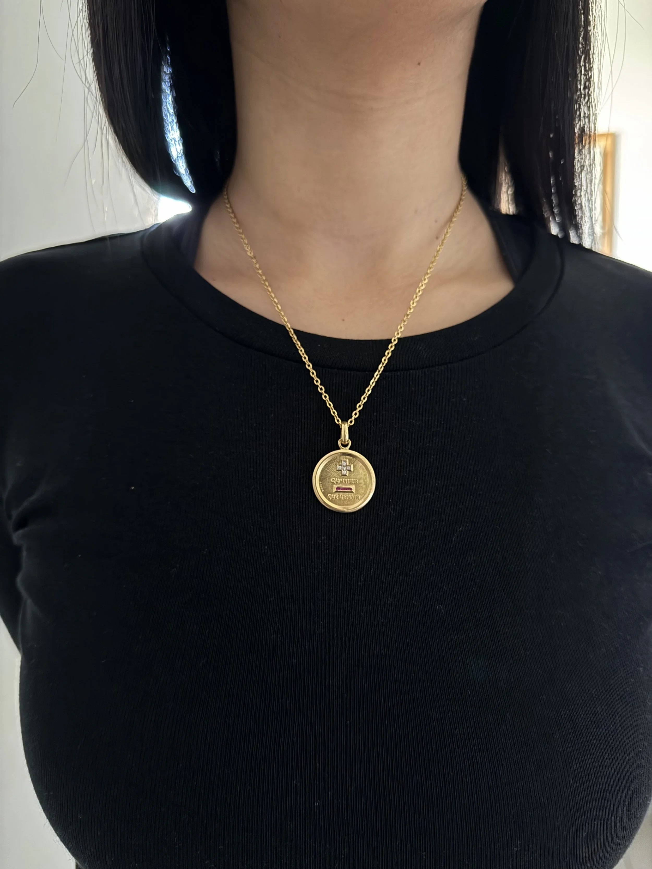 Une personne porte un collier en or avec un pendentif rond. La personne porte un t-shirt noir, et l'image est prise en gros plan sur le cou et le collier.