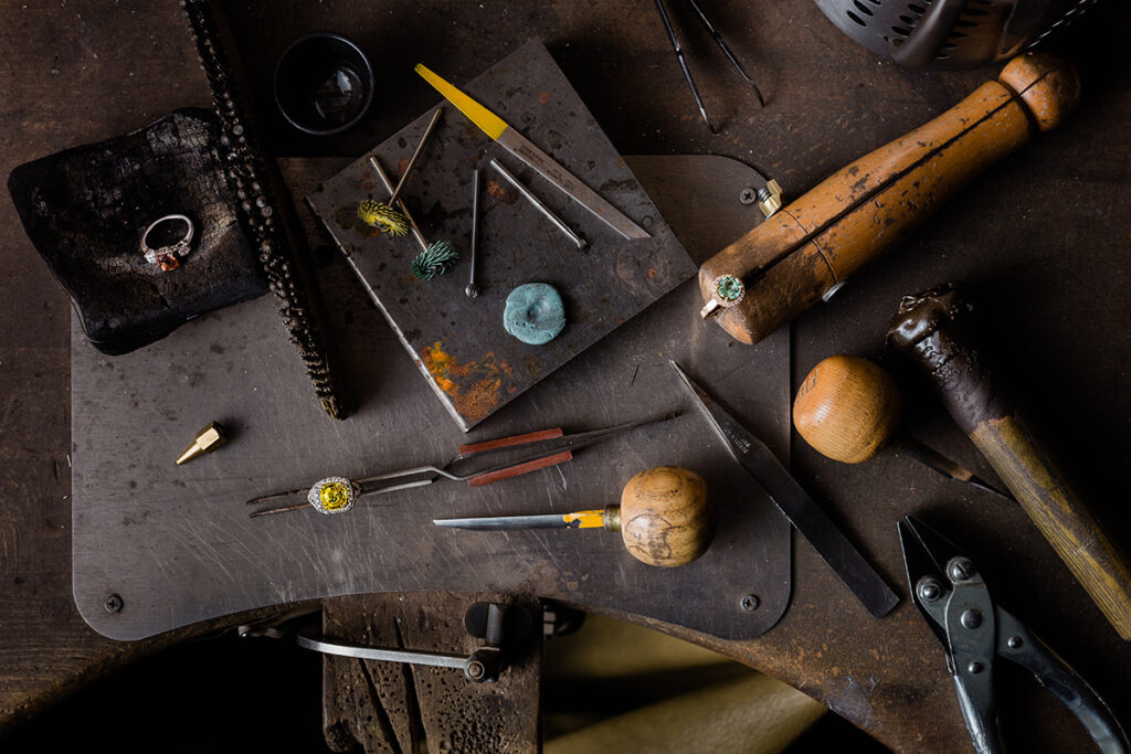 Outils de bijoutier, pince, marteaux, blocs de bois, et bijoux en cours de fabrication sur un établi en métal.