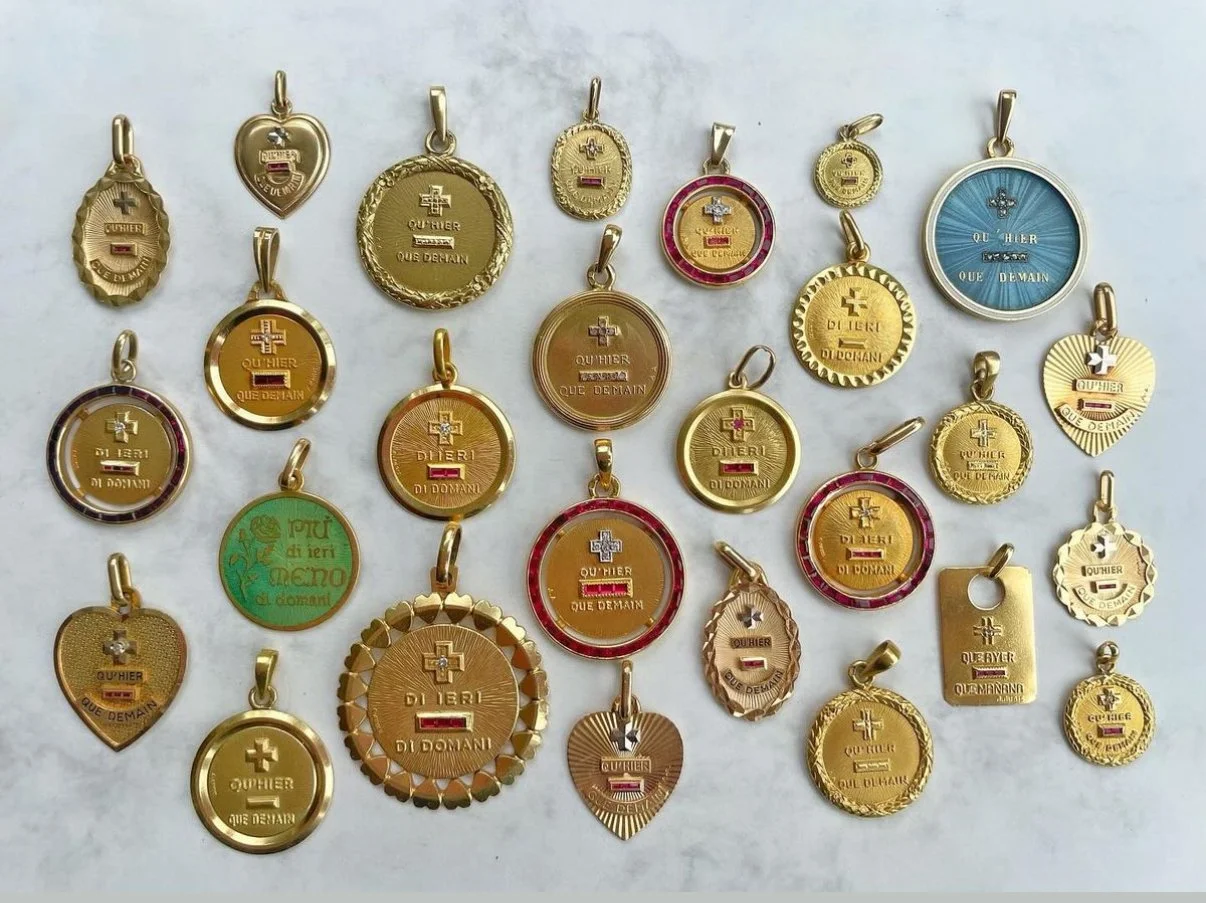 Une collection de pendentifs en métal de différentes formes. Certains sont ronds, d'autres en cœur, en ovales ou en forme de plaque. Tous portent une inscription en français ou en italien, avec des mots tels que « Qu'Hier », « Que Demain » ou « Di Ieri Di Domani ». Certains ont des décorations colorées ou incrustées, d'autres sont simplement en métal doré ou argenté.