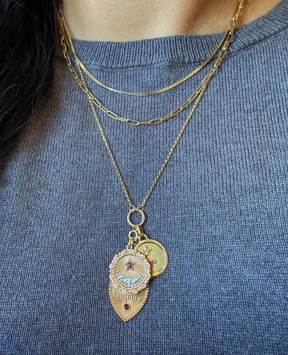 Une personne porte plusieurs colliers en or avec des pendentifs, dont un avec une médaille en forme de bouclier portant l'inscription 'Jouons' et d'autres pièces de monnaie