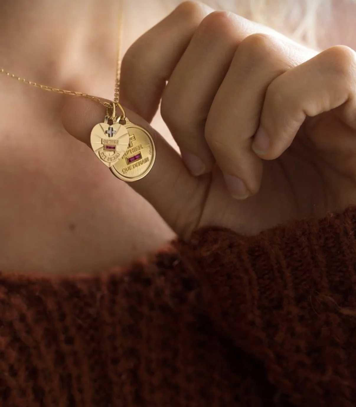 Mains tenant deux pendentifs en forme de pièces de monnaie en or, portant des insignes et des drapeaux, dont un avec la Croix de Lorraine et l'autre avec un drapeau français, porte un pull en laine marron.