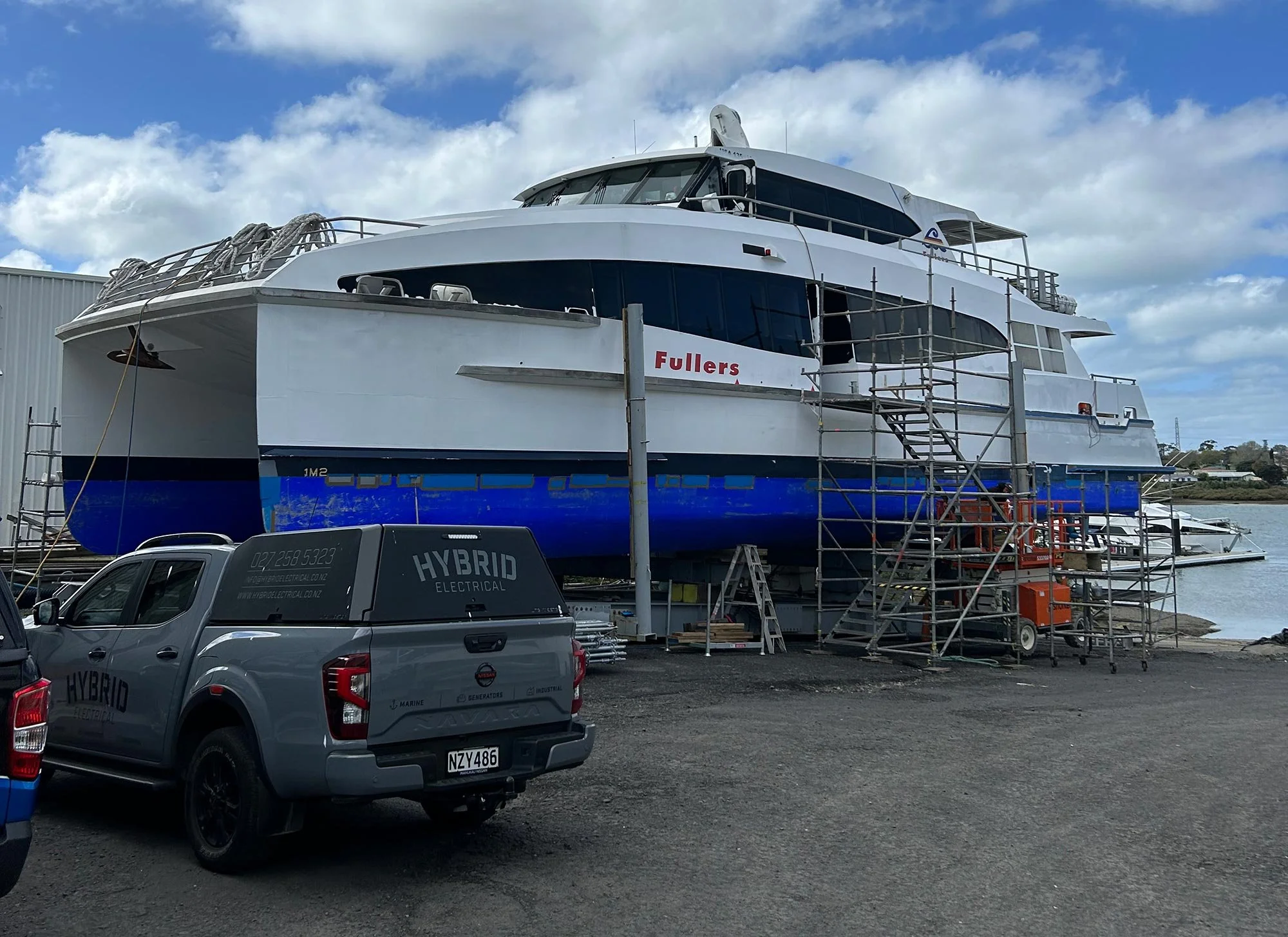 Fullers Ferry docked on land at Orams Marine in Auckland with scaffolding around it, and a grey hybrid electrical work vehicle parked in front.