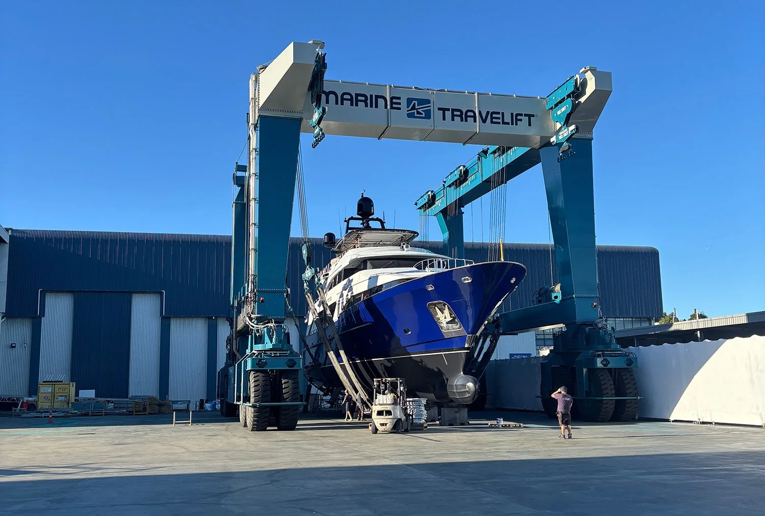 A large super yacht using the marine travelift at Orams Marine Village in Auckland with blue sky overhead and a person on the ground nearby.