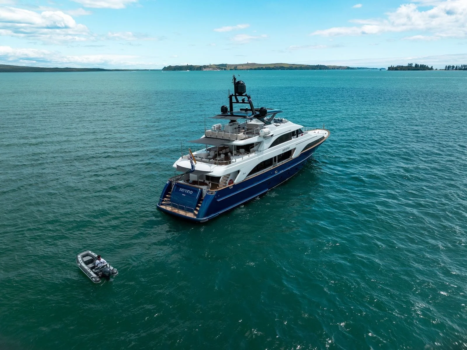 A large white and blue super yacht named 'Sereno' floating in a calm body of water, with a small inflatable boat nearby. The background shows distant land and a partly cloudy sky.