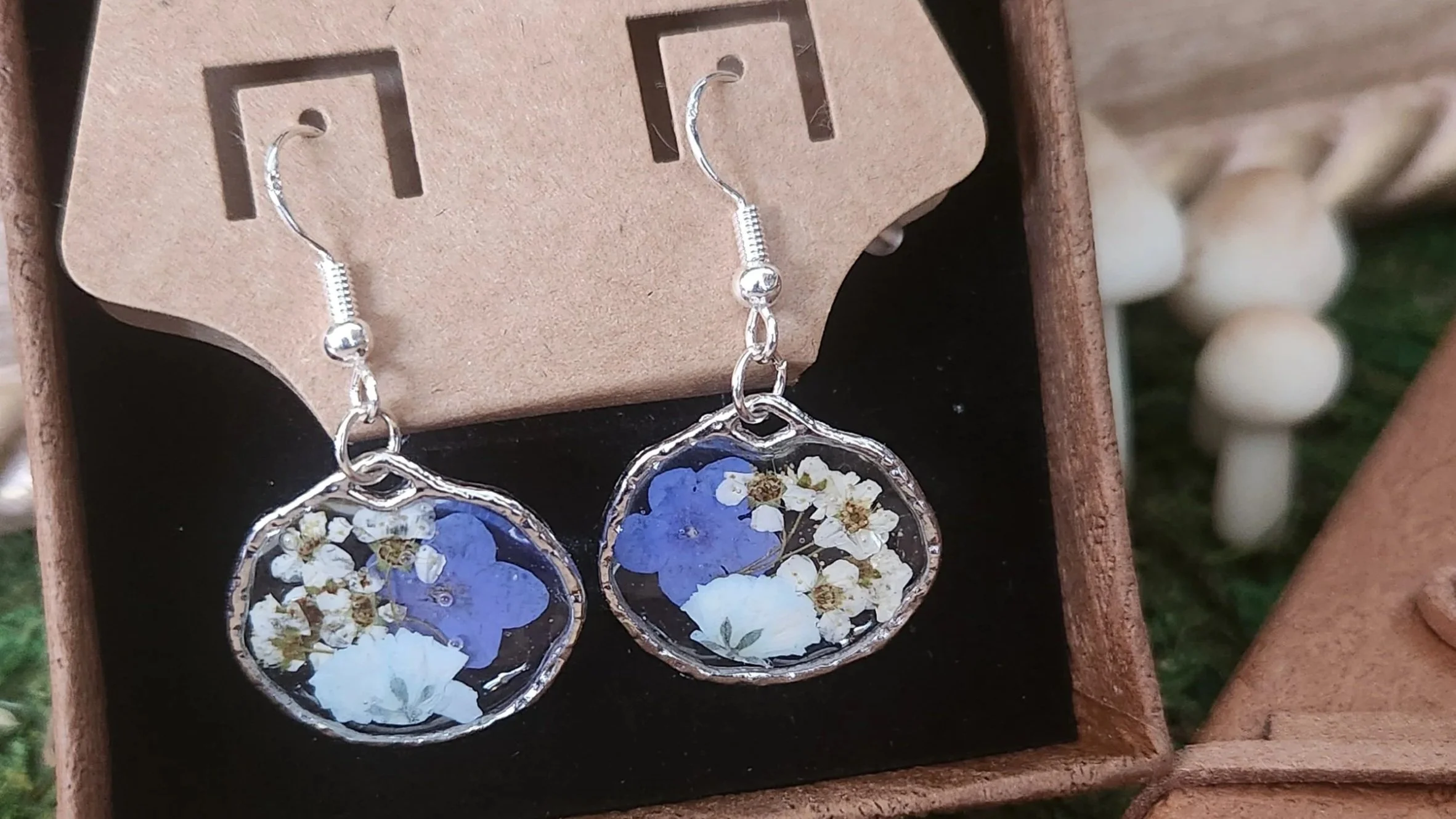 Silver dangle earrings with transparent circular pendants containing blue and white pressed flowers, displayed on a brown jewelry card inside a wooden box.