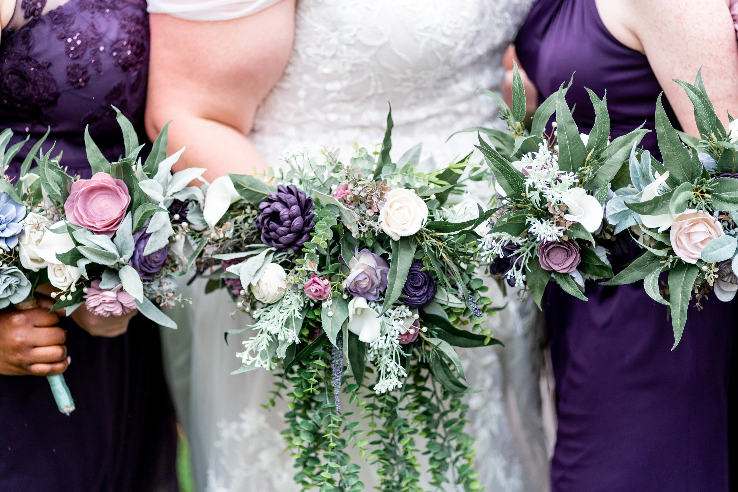 Close-up of three women holding floral wedding bouquets with purple, white, and pink flowers and green foliage.