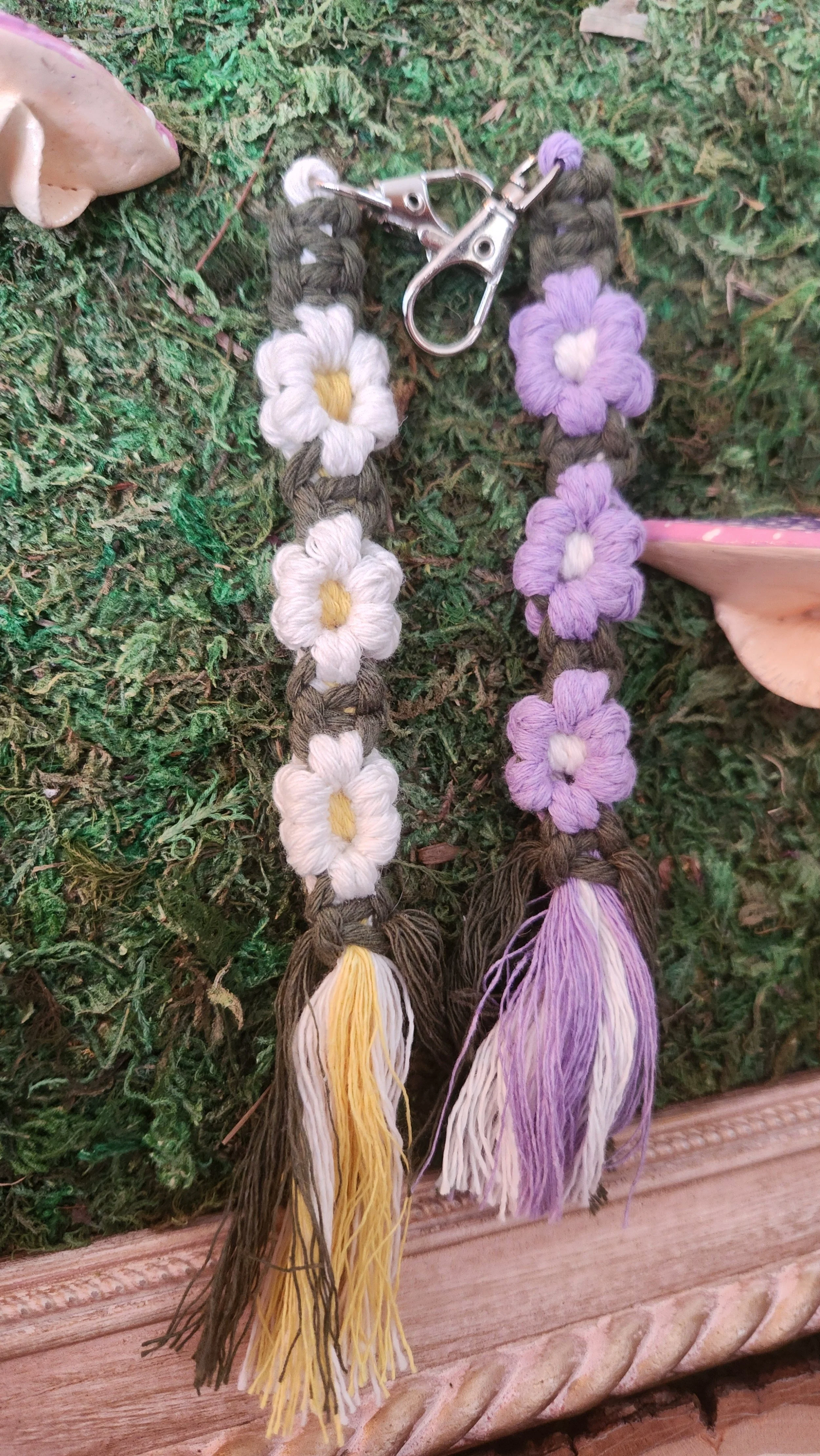 Two handmade yarn keychains with floral designs, one in white and yellow, the other in purple and white, placed on green mossy ground.