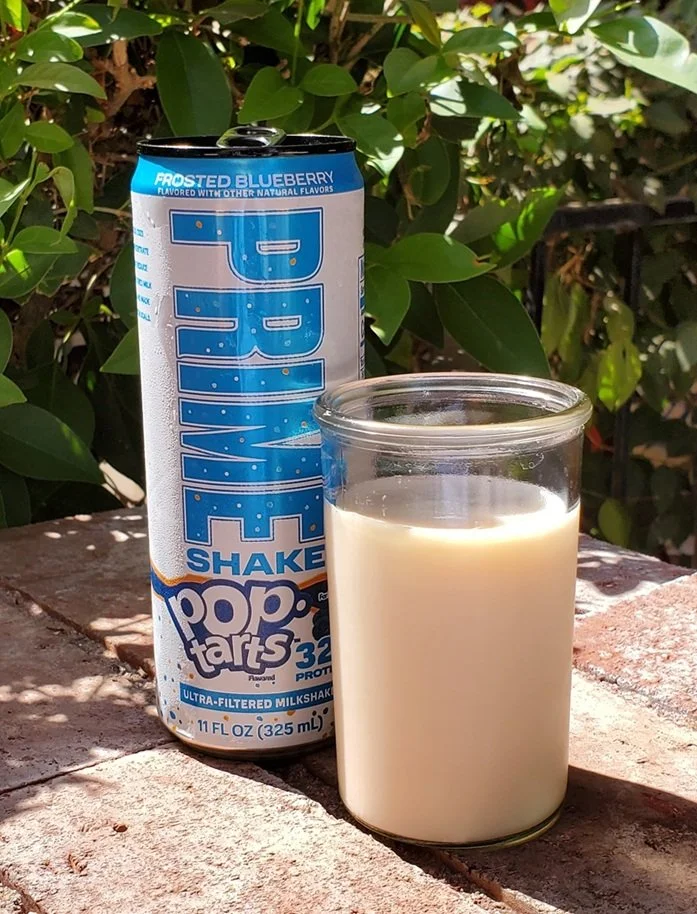 A can of Prime Frosted Blueberry Pop-Tarts Shake stands beside a clear glass filled with the creamy, light-colored milkshake. Both are sitting on a sunlit brick patio surrounded by green foliage.