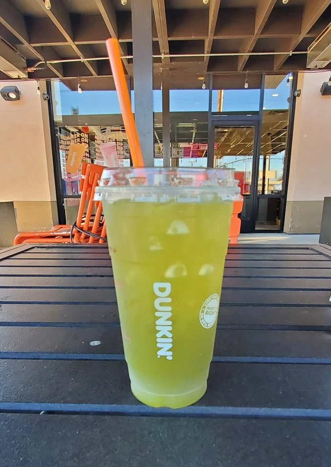 A clear plastic cup of Dunkin’ Green Apple Zero Drink with a straw, sitting on an outdoor table in front of a Dunkin’ entrance.