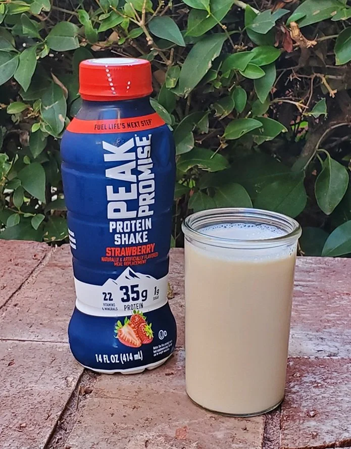 Peak Promise Strawberry protein shake bottle beside a glass of strawberry protein shake outdoors.