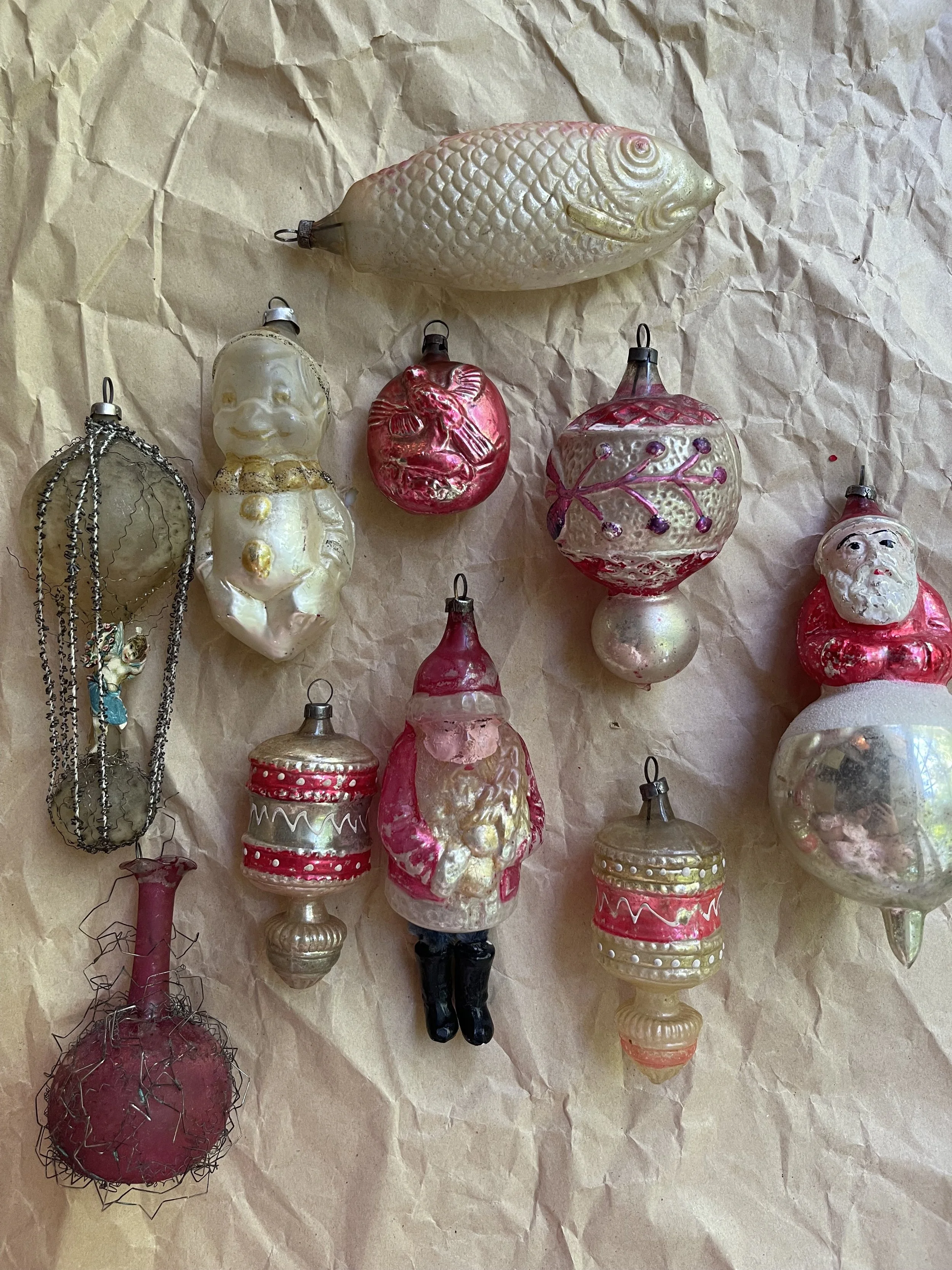 Collection of vintage glass Christmas ornaments on crumpled brown paper, including a fish, a snowman, Santa Claus, decorated balls, and other various themed ornaments.