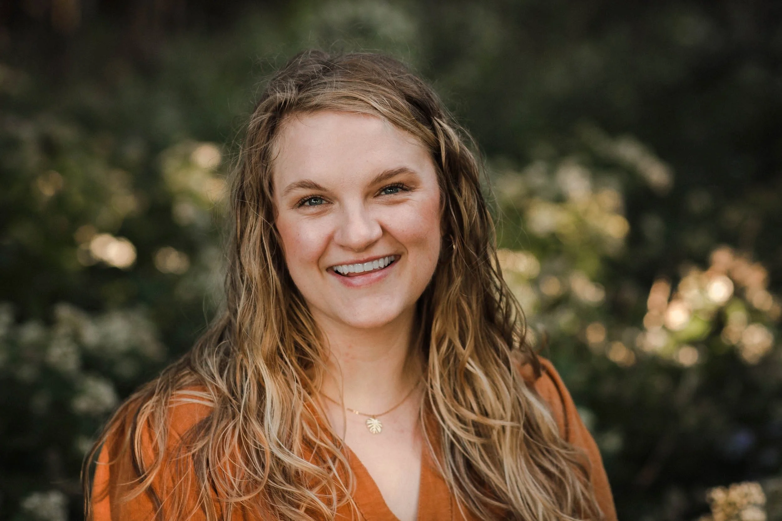 Young woman with long wavy blonde hair smiling outdoors during daytime, wearing a brown top and a gold necklace with a leaf pendant, blurred green foliage background.