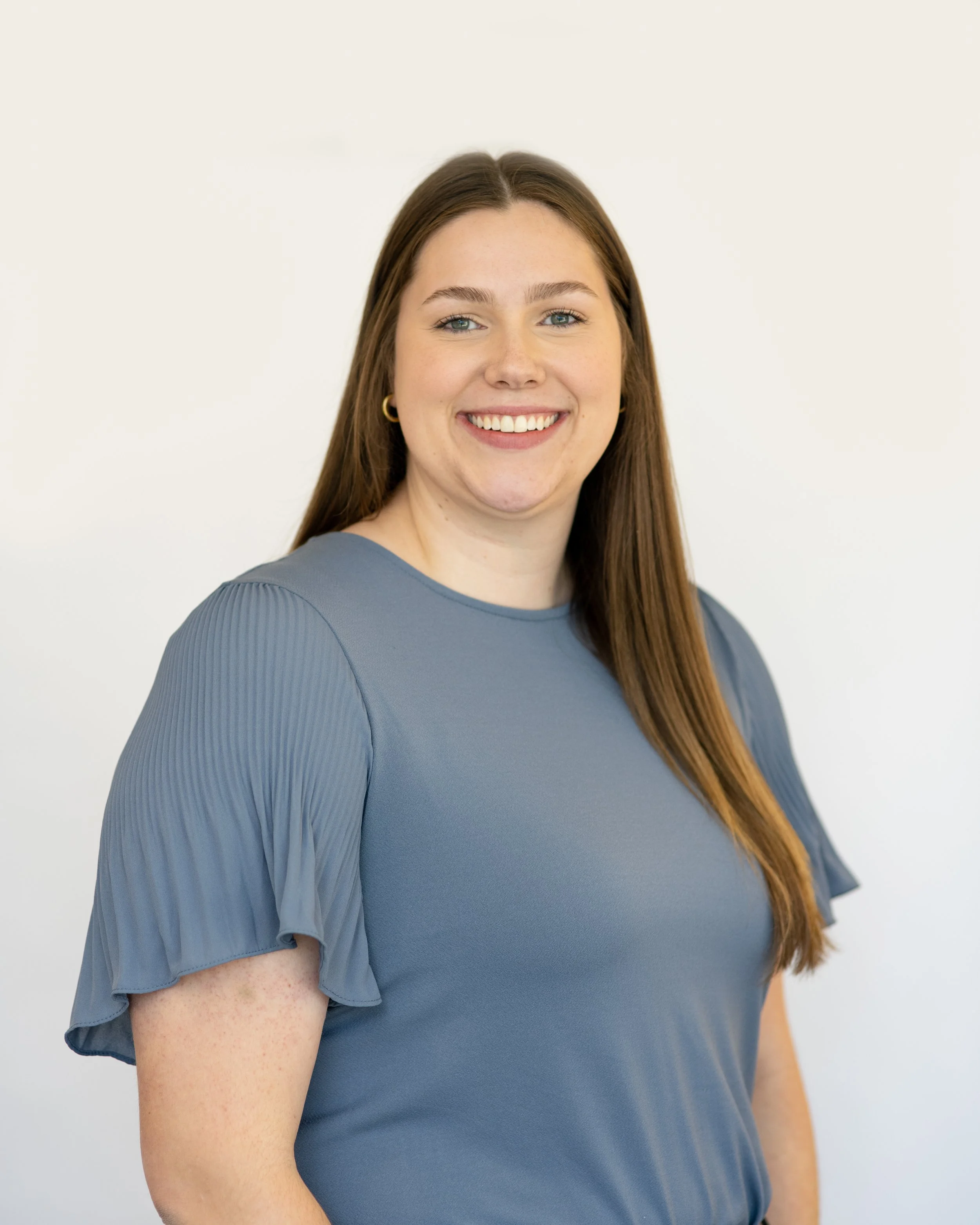 A young woman with long brown hair, wearing a green top and dangling earrings, standing outdoors near a brick wall with greenery in the background.