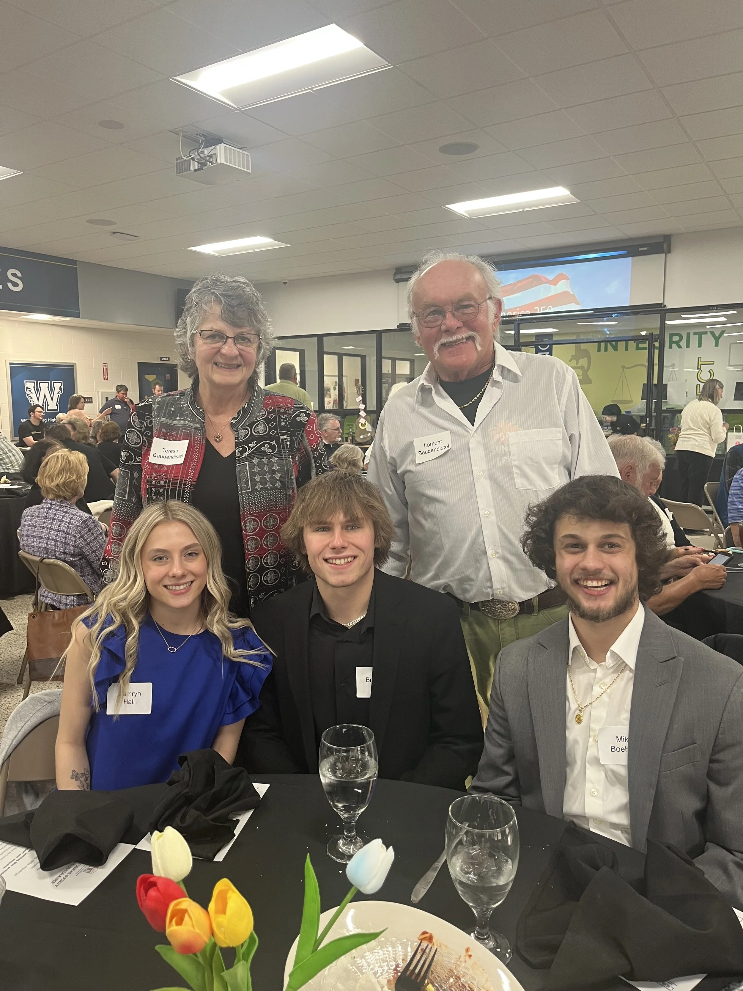 Group of five people attending a formal event in a school cafeteria, with tables, chairs, and other guests visible in the background, posing for a photo at a banquet or celebration.