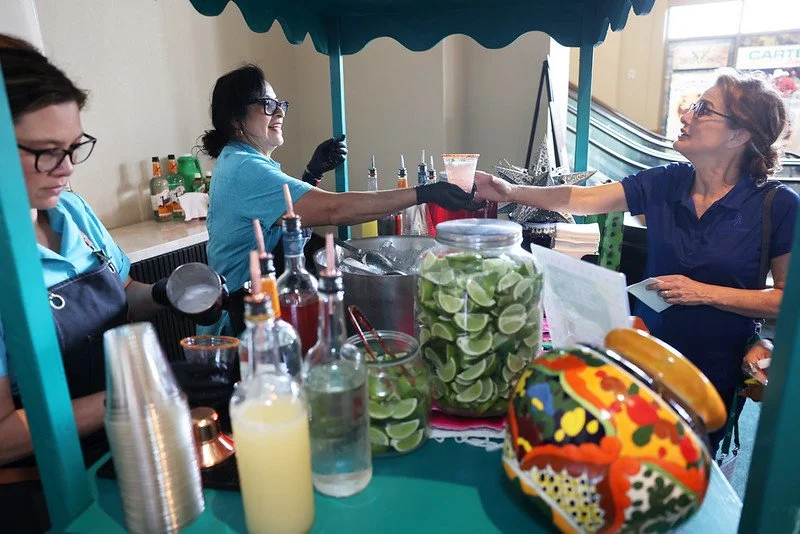A woman in a blue shirt is handing a drink to another woman at a beverage stand with lime slices, bottles, and colorful items.