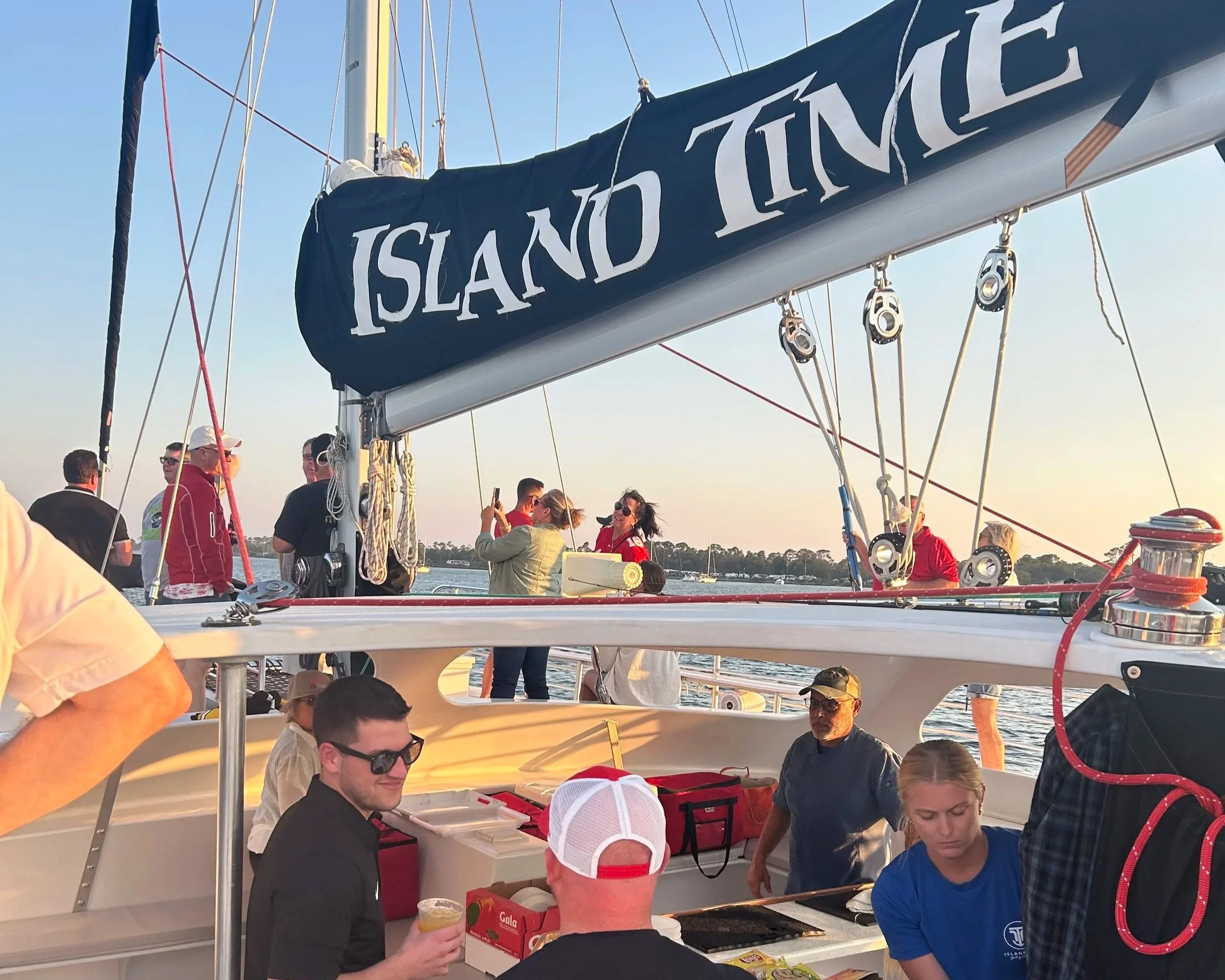People on a sailboat with a large sail labeled 'Island Time' on a body of water during sunset, some taking photos, others socializing and preparing food on the boat.