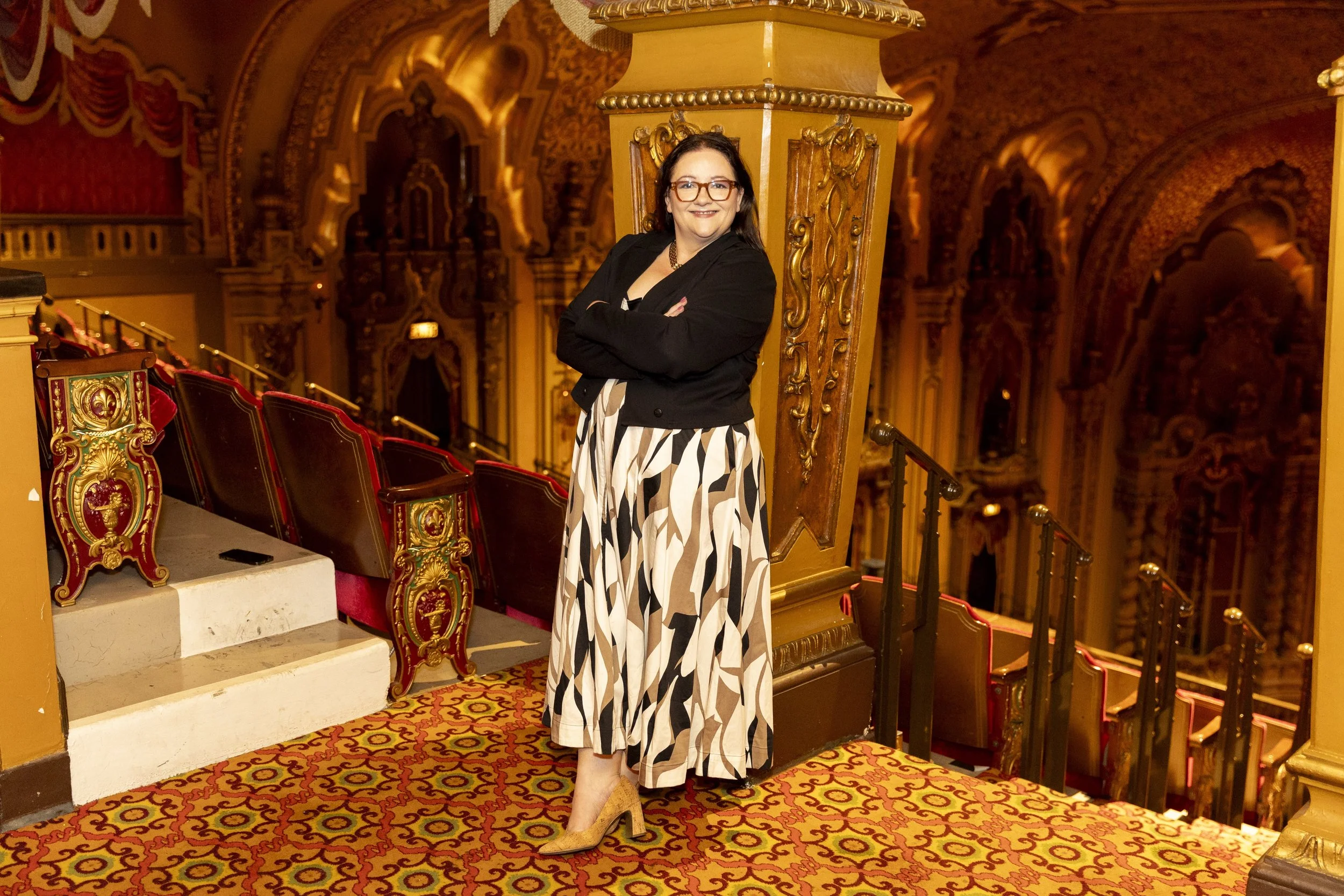 A woman with glasses, dark hair, and a black jacket standing on a theatre balcony with ornate red and gold seats and intricate gold decorations inside a grand opera house.