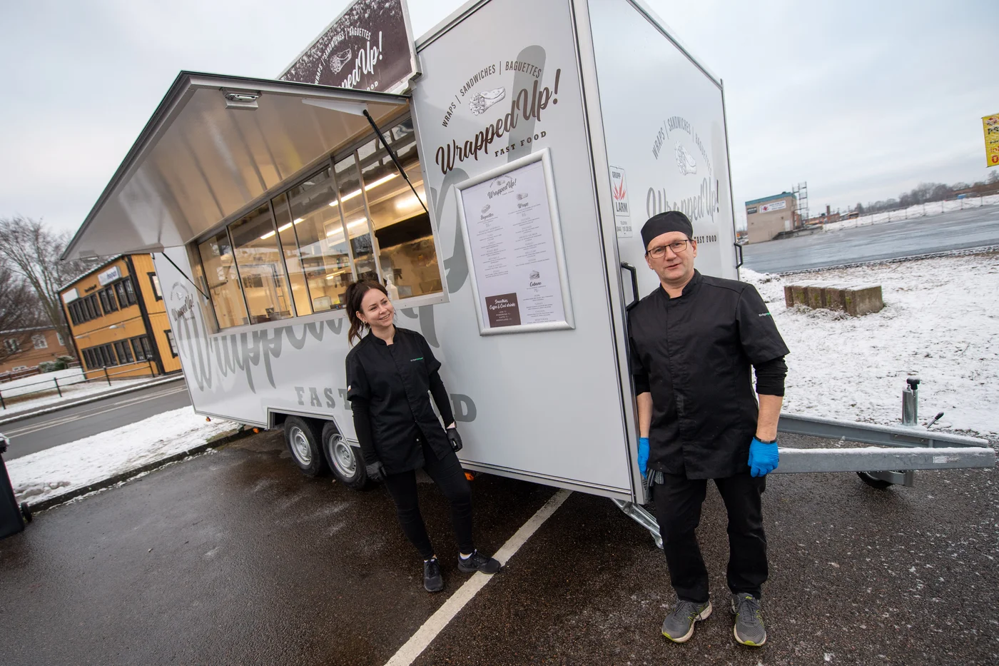 Two food service workers standing outside a food truck titled 'Wrapped Up!'. One is a woman with dark hair, smiling, and the other is a man with glasses and a black beanie, wearing blue gloves. Snow is on the ground.