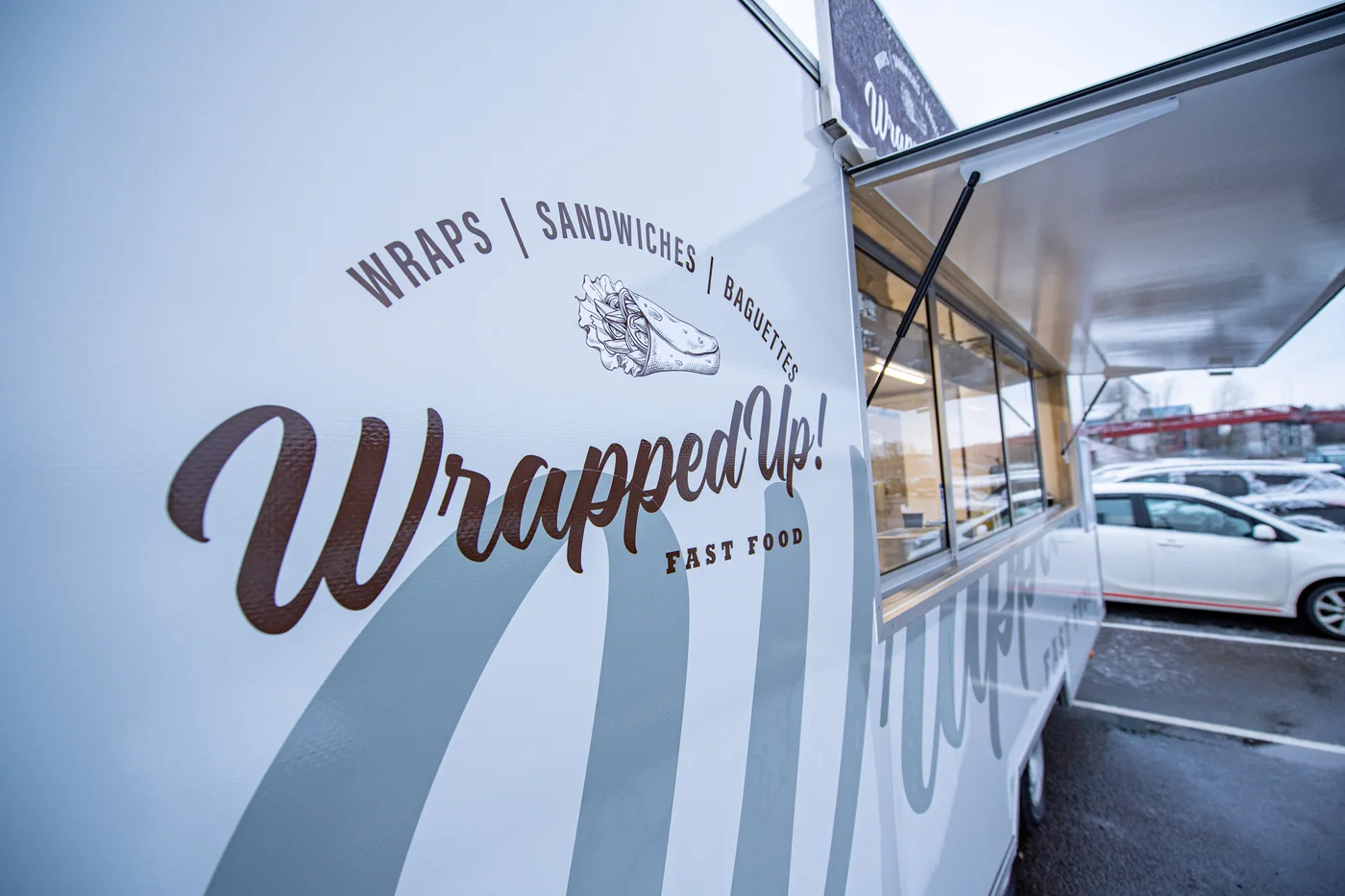 Side view of a white food truck with a logo that reads "Wrapped Up!" and lists menu items: wraps, sandwiches, baguettes, and fast food.