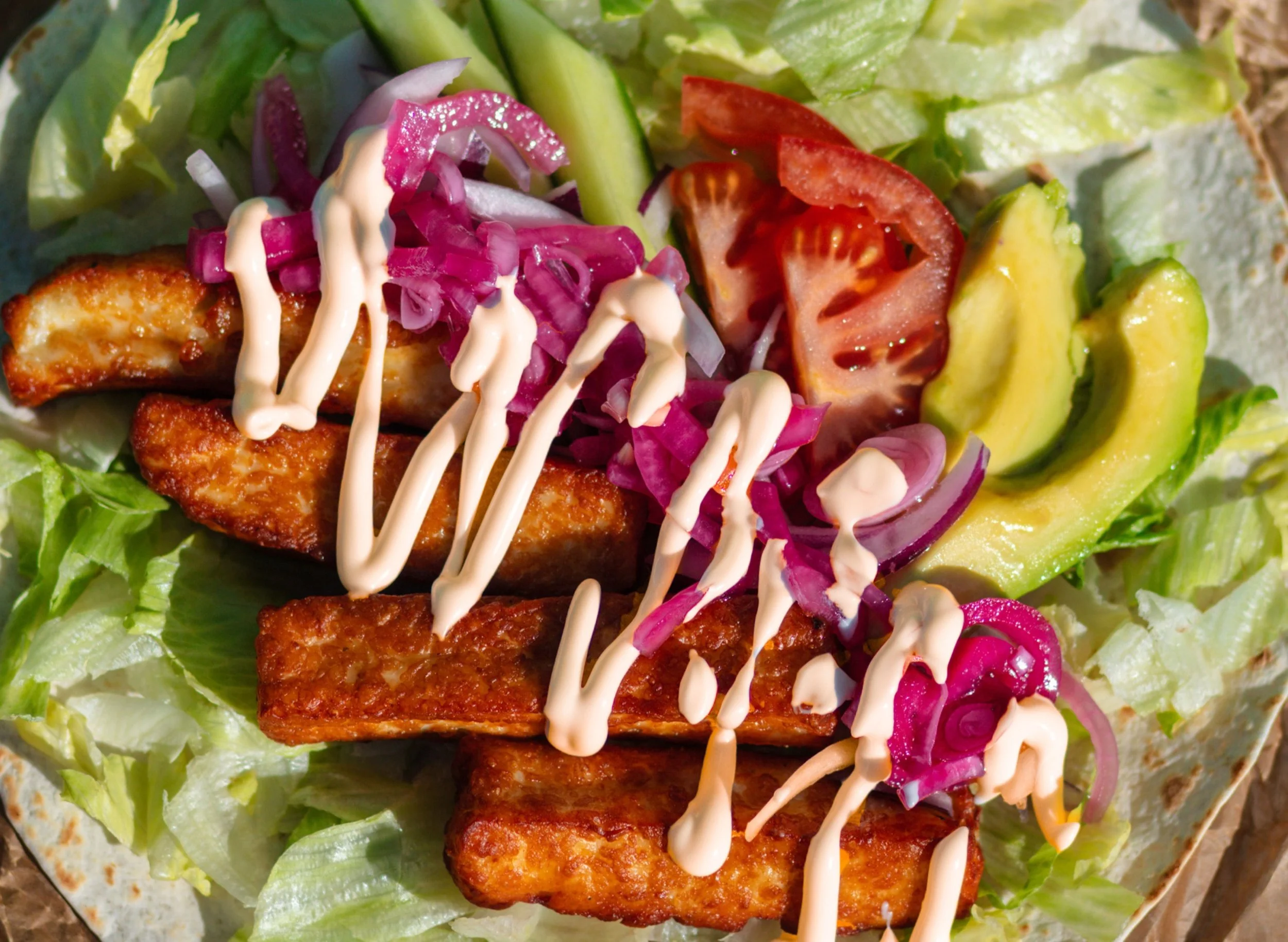 Close-up of a salad with grilled chicken strips, avocado slices, tomato wedges, cabbage, and pickled onions, topped with a drizzle of creamy dressing.
