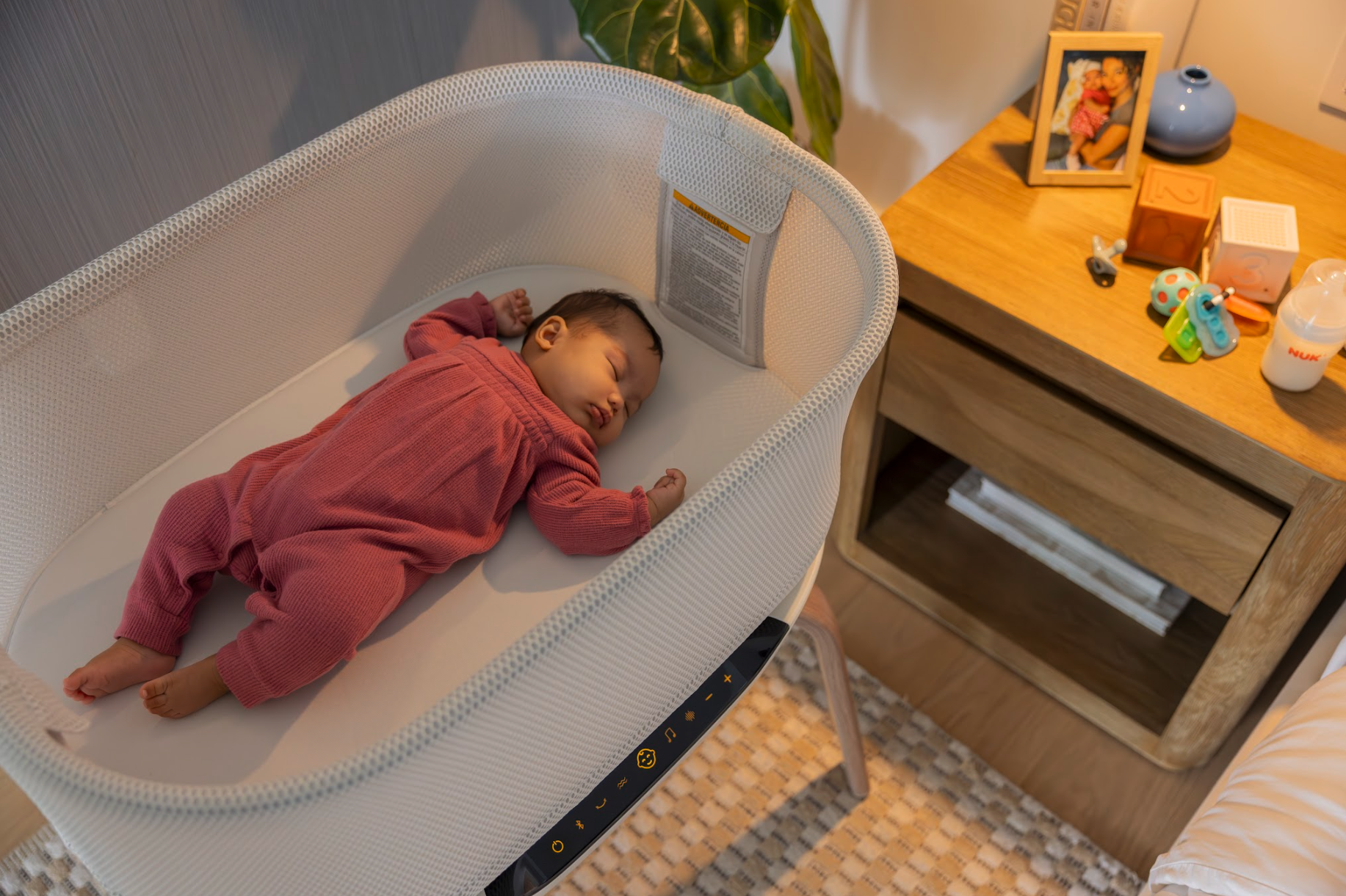 A baby sleeping in a white bassinet next to a wooden nightstand with family photos and baby items.