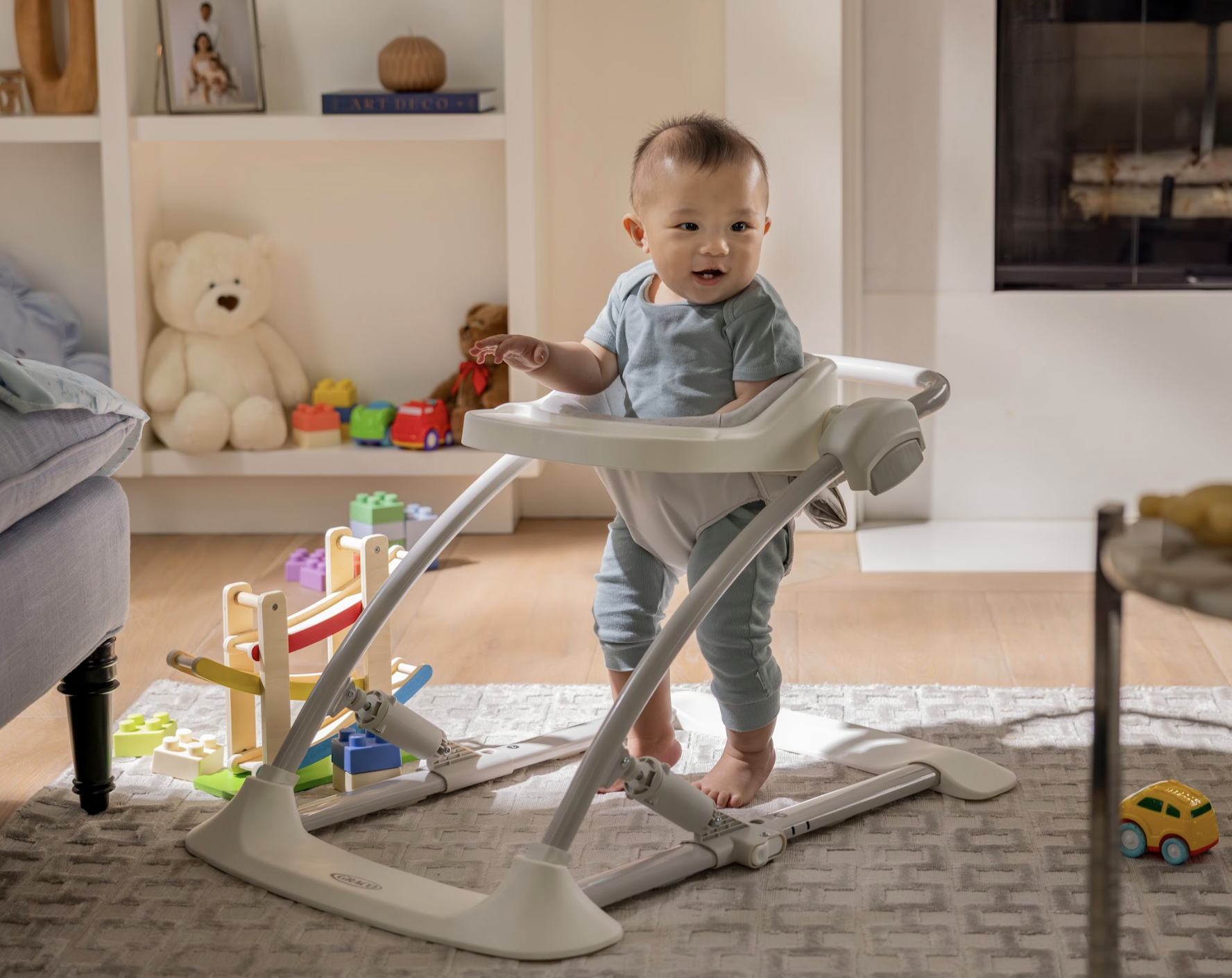 A toddler standing in a white baby walker in a cozy living room with toys and stuffed animals in the background.