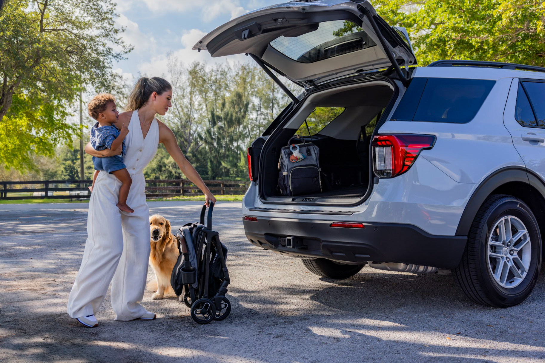 A woman loading a backpack into the trunk of an SUV with a child in her arms and a dog standing beside her in a park-like setting.