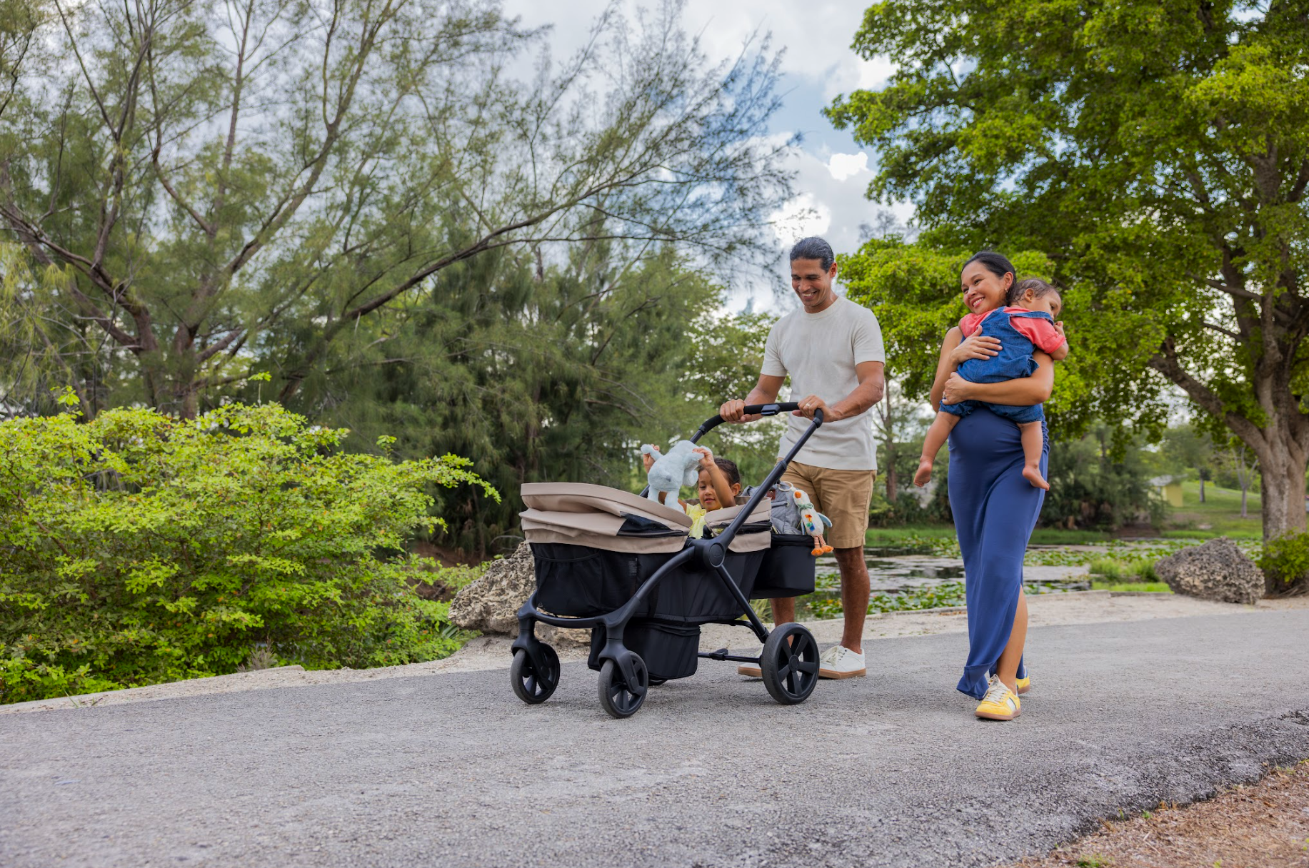 A family enjoying a walk in the park. The father is pushing a stroller with a young child, the mother is carrying another child, and they are all smiling amidst lush green trees and a pond in the background.