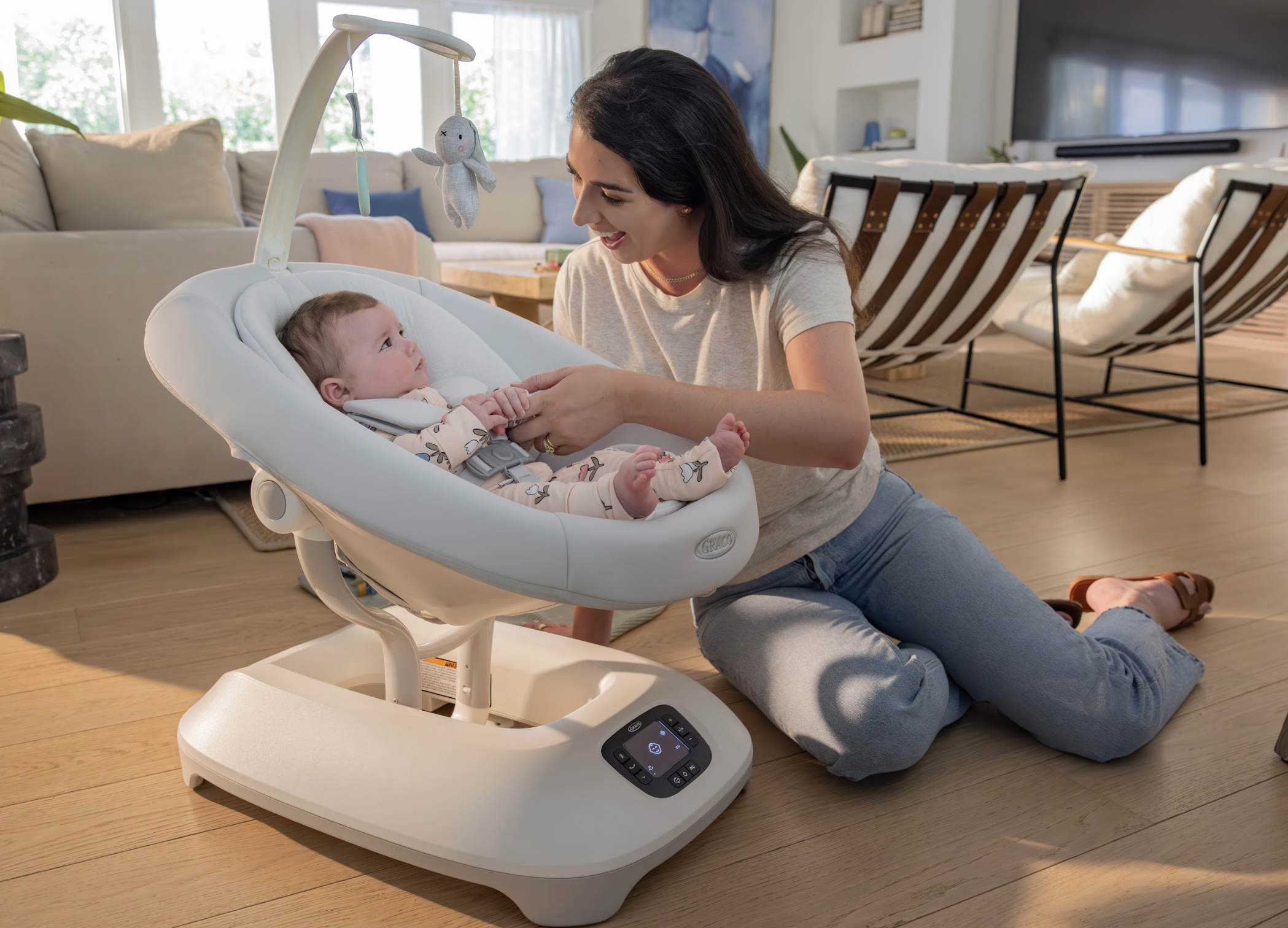 A woman kneeling on the floor playing with a baby in a baby swing in a living room.