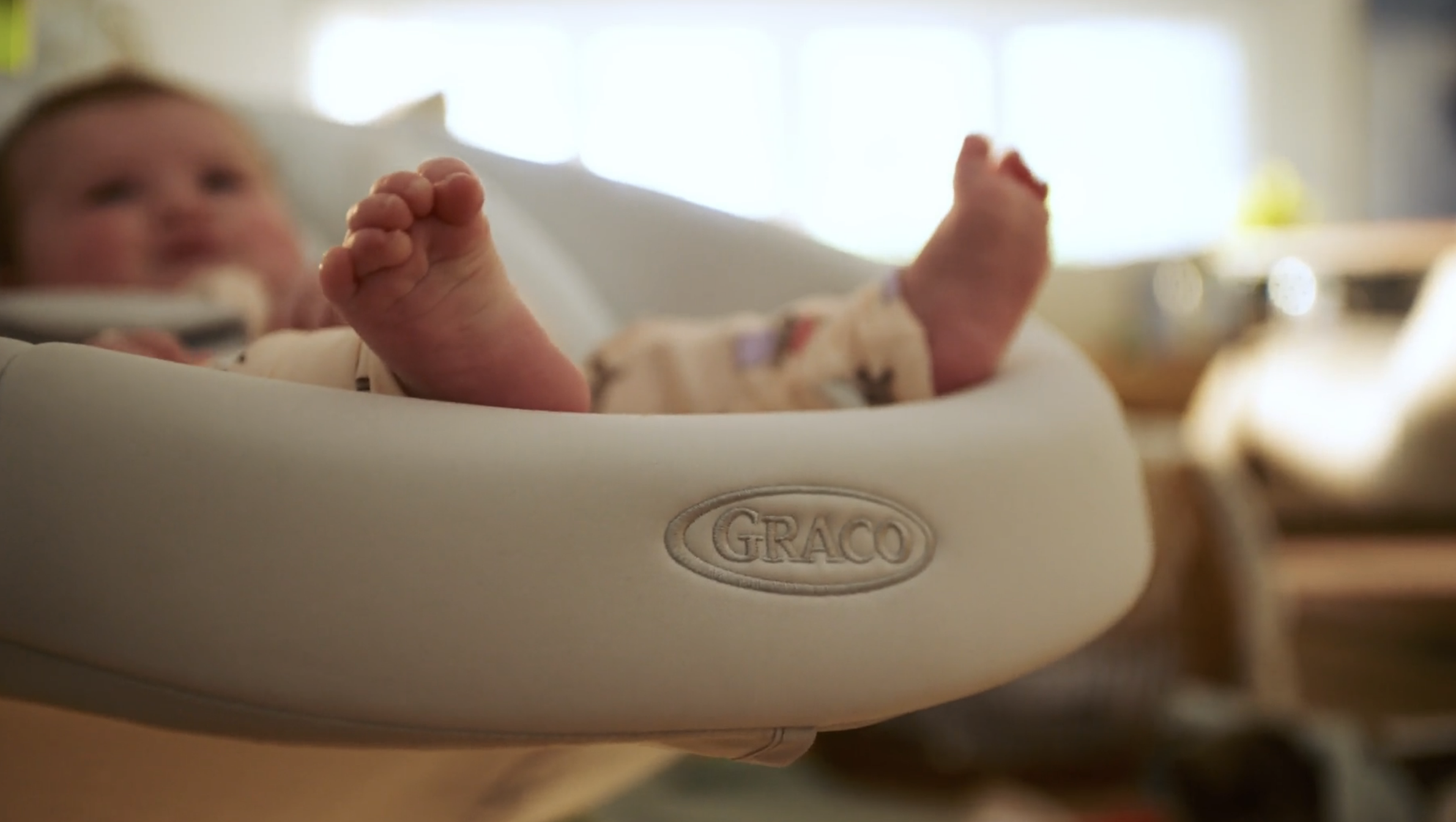 Close-up of a baby's feet resting in a Graco baby swing, with the baby's face blurred in the background.