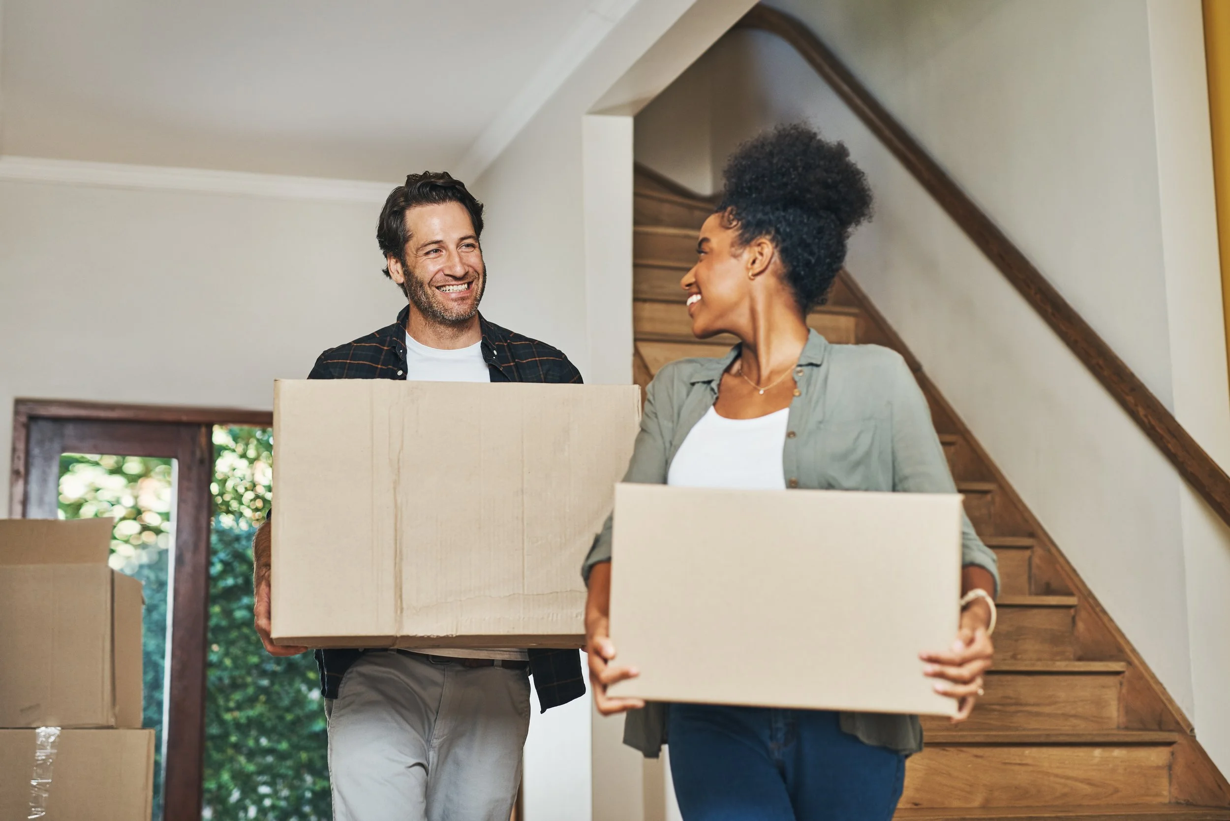 A man and woman carrying cardboard boxes inside a house, smiling at each other, with a staircase and a door with glass windows in the background.