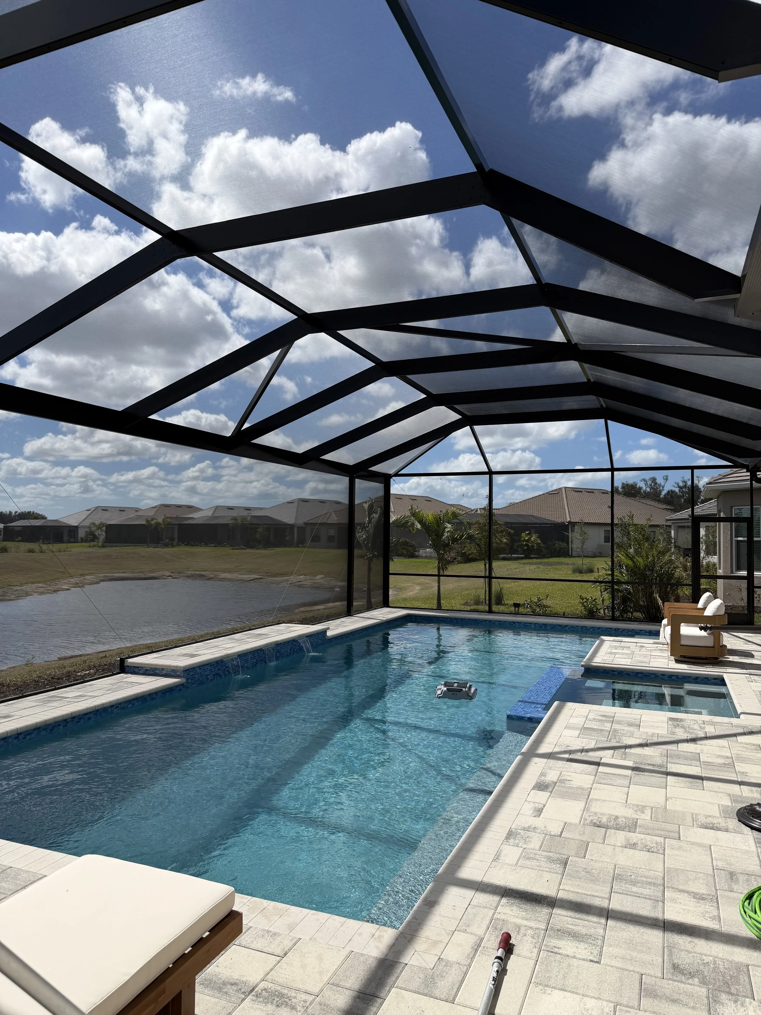 A backyard enclosed patio with a swimming pool, screened with a metal frame and fine mesh, under a partly cloudy sky during daytime. There are poolside chairs and a small pond beyond the screened enclosure.