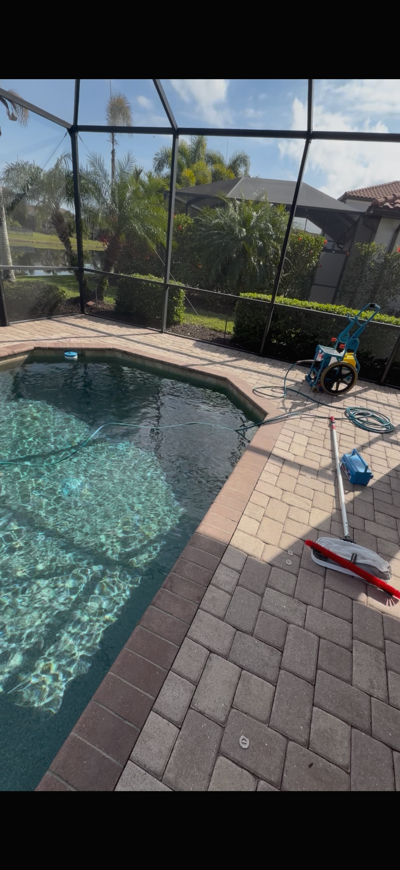 Pool enclosure with swimming pool, pool cleaning equipment, and patio pavers, surrounded by lush greenery and trees.