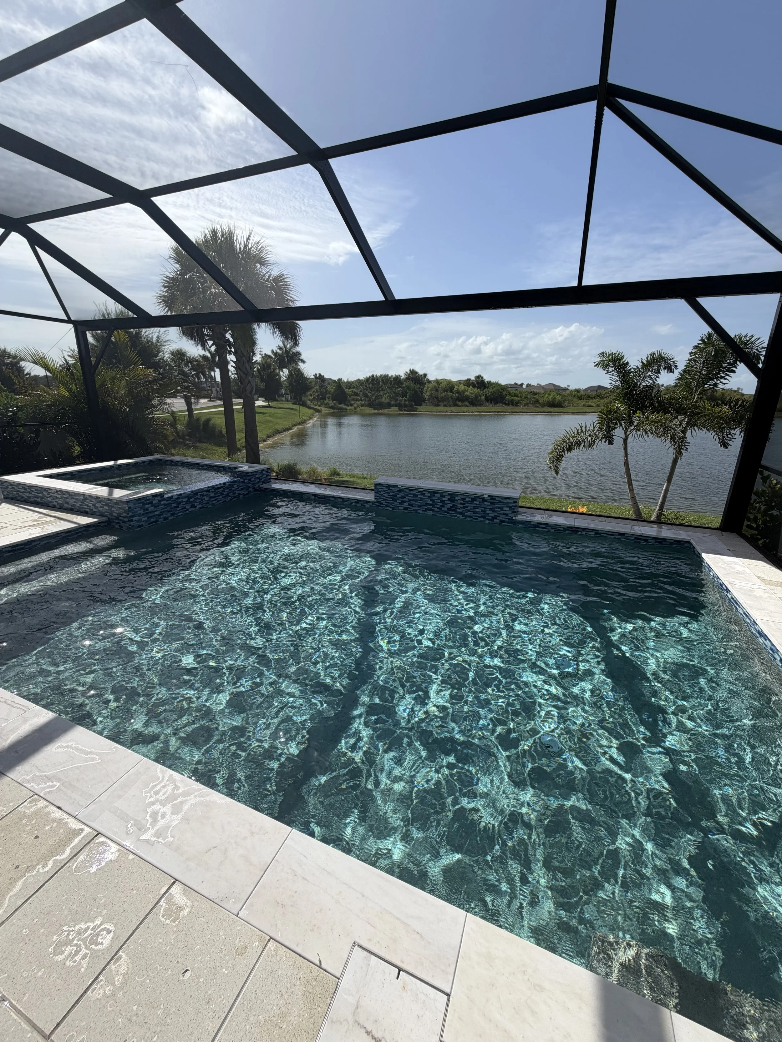 An outdoor swimming pool and hot tub with a brick border on a white patio, surrounded by pool float toys, patio furniture, and garden hoses.